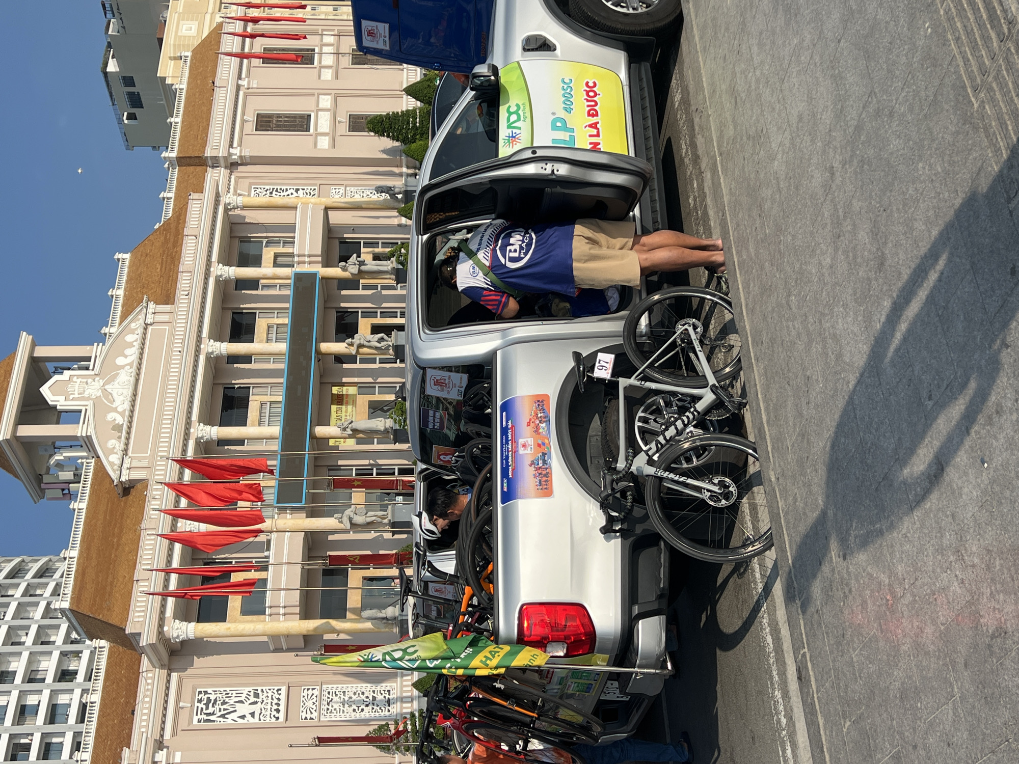 The image captures a scene in Nha Trang, Vietnam, featuring a light-colored pickup truck loaded with bicycles. A man wearing a helmet and a blue and white cycling jersey with the text "BM PLACE" is leaning into the driver's side of the truck. The truck has various stickers and logos on it, including one for "LP 400SC." The bicycles, some with the brand name "FACTOR," are mounted on the truck bed. The backdrop shows a building with flags and statues, suggesting it might be a government building or a historical landmark. Red flags are prominently displayed in front of the building. The image appears to be taken during the day, with bright sunlight casting shadows on the pavement.