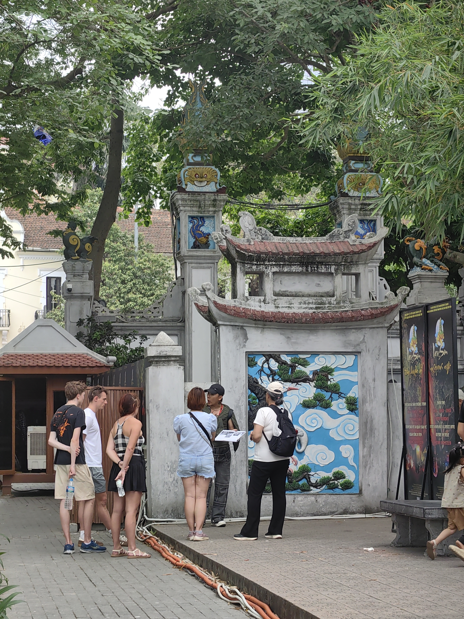 The image shows a group of tourists gathered around a guide or local person, in front of an ornate stone gate. The group consists of two men and a woman, listening to the guide in a black baseball cap. There is a woman in a white T-shirt with a black backpack. A child is partially seen on the right. The gate features elaborate carvings, including a blue and white cloud-and-pine design on the central panel. Posters are standing beside the gate. The paved ground has exposed orange and white cables. The presence of trees suggests it may be in the city of Hanoi. Based on the poster with Vietnamese text, this may be related to the Lac Son Show.