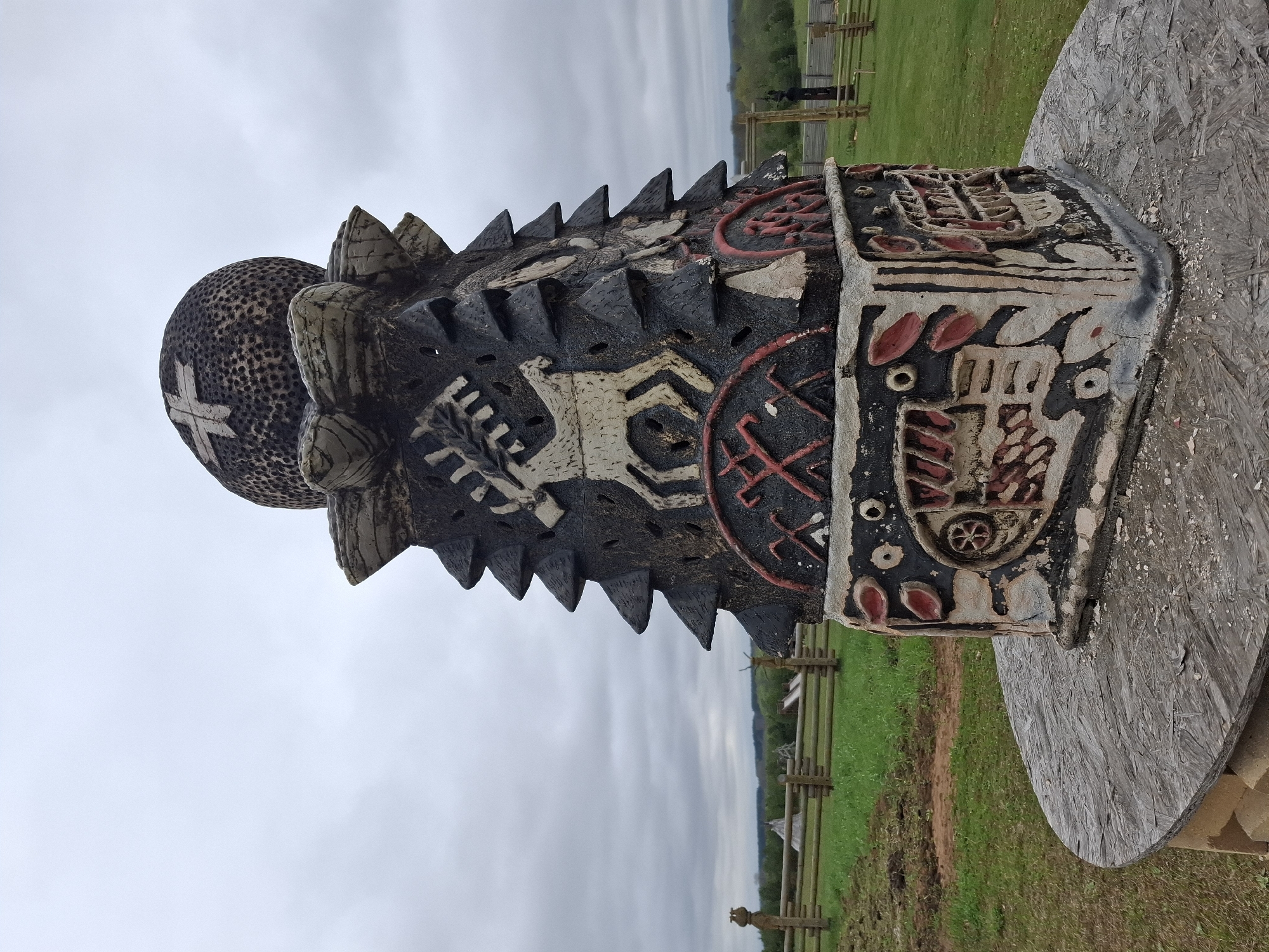 A carved wooden totem pole rests on a weathered circular wooden platform outdoors in a grassy area. The sky is overcast. The totem pole is decorated with geometric shapes, animals, and other designs, painted in black, white, and red. The top of the pole features a textured sphere with a raised cross. The background includes a wooden fence and rolling green hills. Адам, Russia is not definitively discernible in the image.