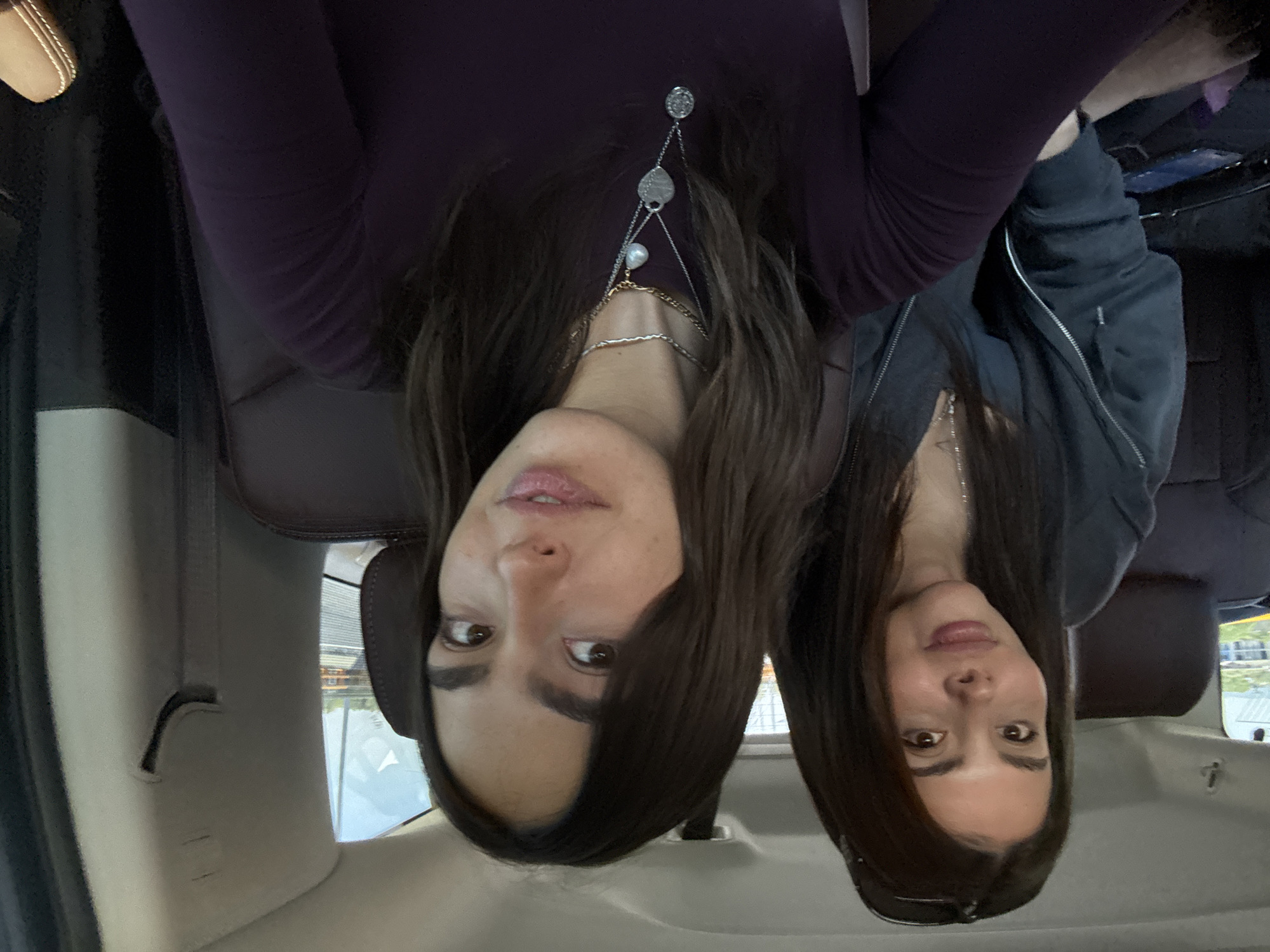 The image displays two women seated inside a vehicle, presented in an inverted (upside down) orientation.
On the left side of the frame, a woman with long, dark brown hair is visible, wearing a dark purple long-sleeved top. She has a prominent silver-toned necklace featuring multiple delicate chains, a white pearl, a round textured charm, and a heart-shaped pendant. Her lips are slightly parted, and her brown eyes are directed towards the viewer.
To the right, another woman with similar long, dark hair is present. She is dressed in a dark grey zippered jacket or hoodie, and sunglasses are partially visible resting on her head. Her expression is calm, with a subtle smile.
The setting is clearly the interior of a car or SUV, evidenced by the dark leather seats, headrests, and car trim visible around the subjects. Through the vehicle's windows, a blurred outdoor background is visible, suggesting a daytime scene in an urban or suburban environment. The women appear to be posing for a casual photo or selfie. There is no discernible text within the image. The natural lighting indicates it is daytime. The provided context indicates the location is Mexico City, Mexico.
