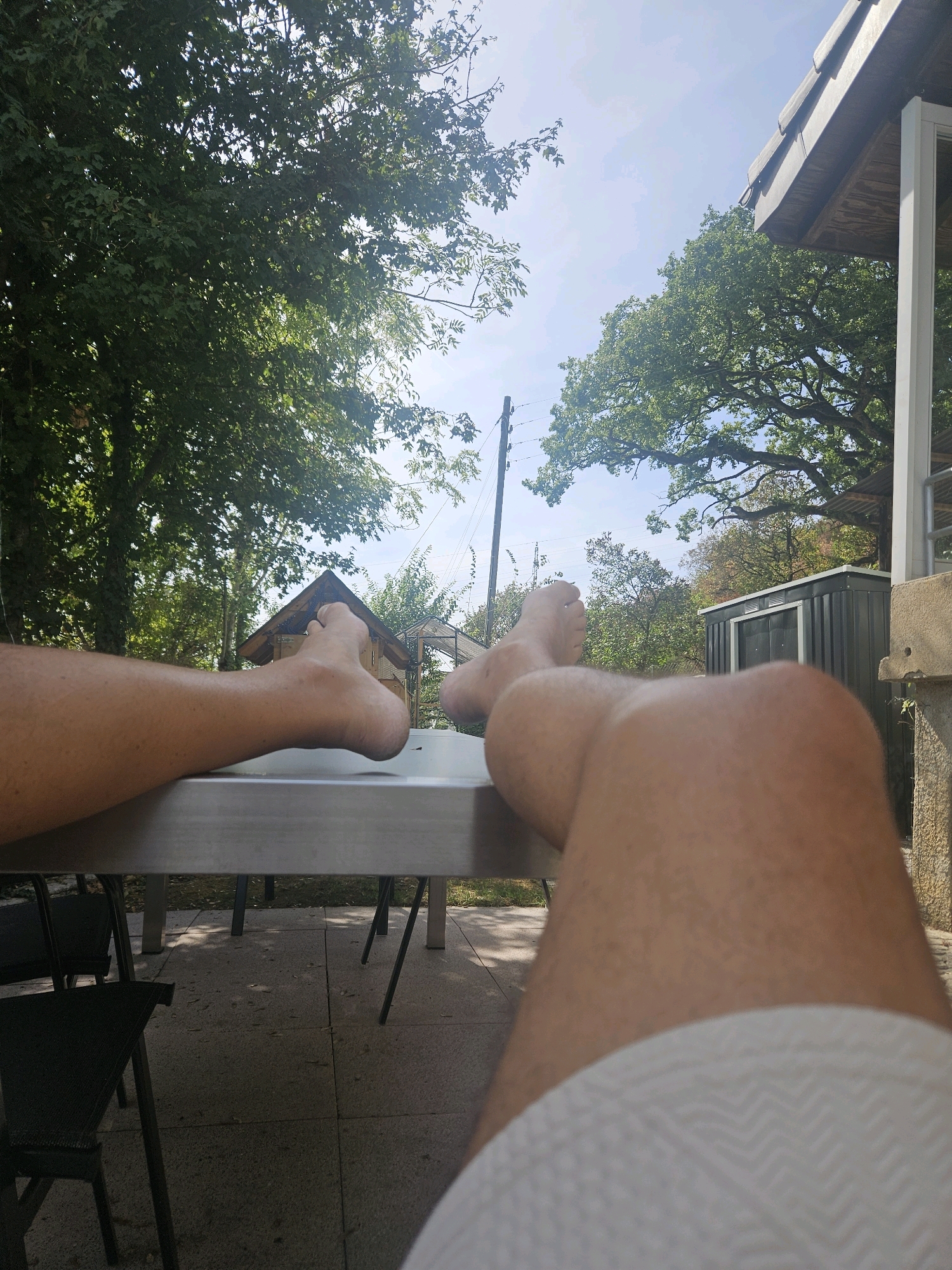 The image shows a person's legs and feet resting on a metallic table outdoors, with a blurred background of trees and a building under a bright sky. The perspective is from the person's viewpoint, looking down at their own legs. The surroundings suggest a relaxed outdoor setting, possibly a patio or garden area with a small structure in the distance. The lighting indicates it is daytime. There are no other people or specific identifiable landmarks that would indicate the location in Cornaux, Switzerland.