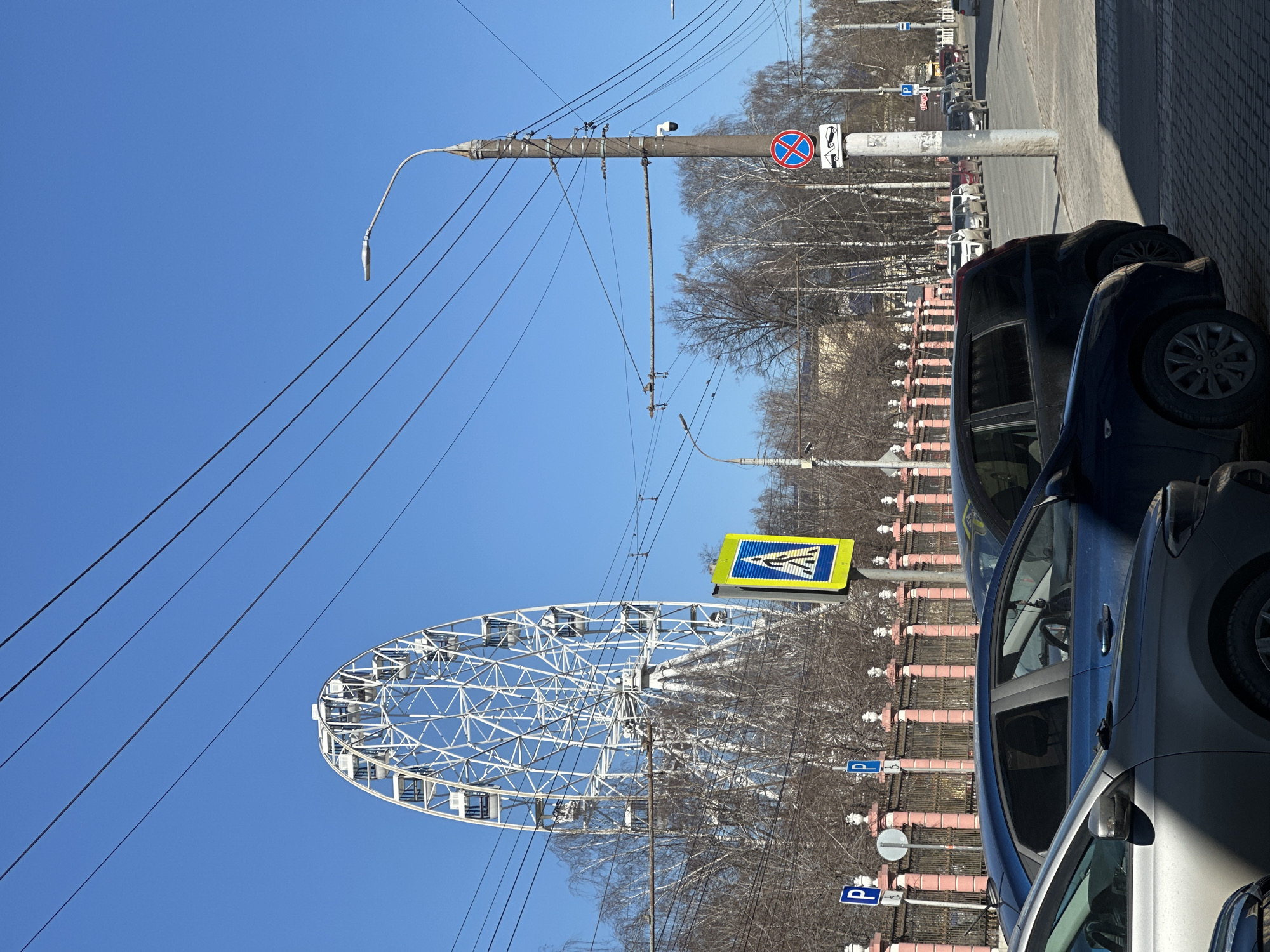 The image shows a street scene in Postol, Russia, featuring a Ferris wheel. The Ferris wheel is white and located on the left side of the image, with a clear blue sky in the background. Overhead power lines stretch across the top left of the image. The right side of the image shows several parked cars along a street with a sidewalk. A pink and white fence separates the sidewalk from a line of trees. Street signs are visible, including a "No Stopping" sign and a "Pedestrian Crossing" sign. Parking signs are also located behind the cars. The image is taken during the daytime with clear sunlight.