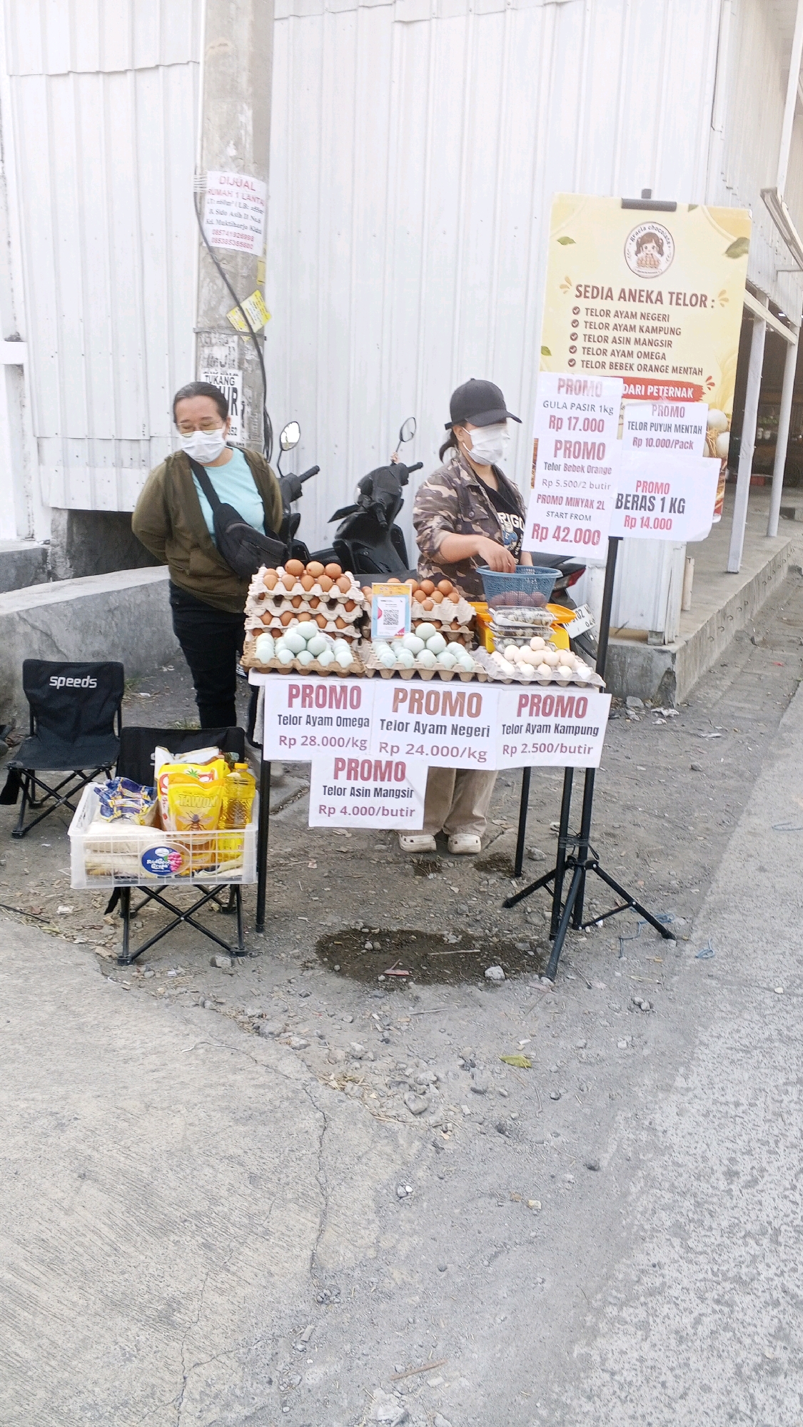 The image shows an outdoor street scene in Semarang, Indonesia, where two women are selling eggs and other food items from a temporary stall. The stall is set up on the side of a street, with a white corrugated metal wall in the background. There are several types of eggs displayed on tables, including chicken eggs and duck eggs. Signs indicate "PROMO" (promotion) prices for various items. One woman, wearing a green jacket and a light blue t-shirt, stands to the left, facing the camera. The other woman, wearing a camouflage jacket and a black cap, is on the right, attending to the stall. A motorcycle is parked behind the women. In the foreground, there are two folding chairs, one with the "Speeds" logo, and a plastic crate containing bags of groceries, including cooking oil and what appears to be rice. The ground is paved with concrete and some gravel.