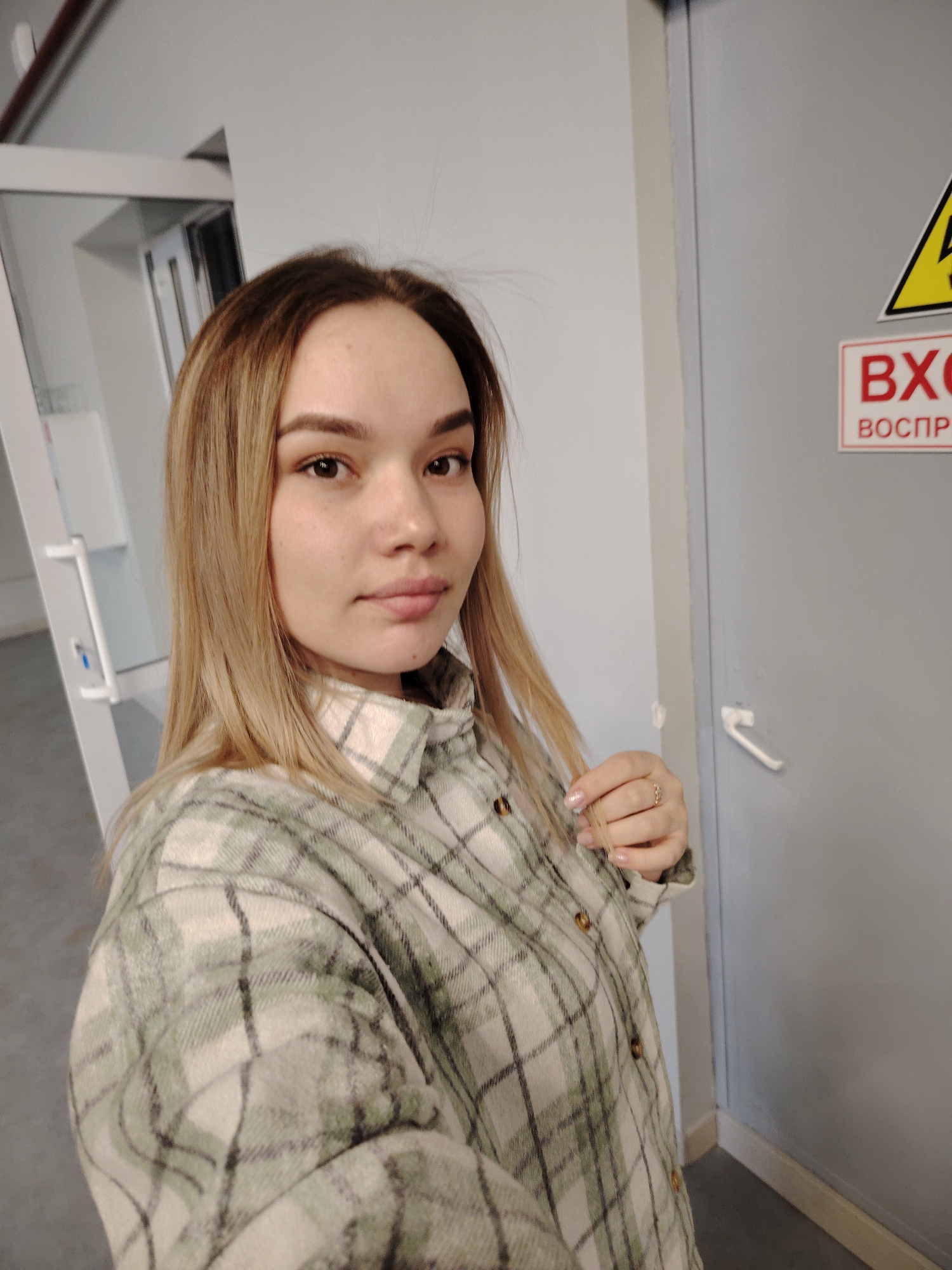 A young woman with long, straight, light blonde or brown hair is captured in a selfie taken indoors. She has light skin, dark eyes, and a neutral, calm expression, looking directly at the camera. She is wearing an oversized, light-colored flannel shirt with a plaid pattern featuring white/cream, green, and dark gray checks. A golden ring is visible on her right ring finger as she gently touches a strand of her hair.
The background features a plain gray door on the right with a white handle. Attached to this door are two signs: a yellow triangular warning sign with a black border and a black lightning bolt symbol, indicating a hazard, and below it, a red rectangular sign with white text. The visible text on the red sign reads "ВХОД" (Entry/Entrance) and "ВОСПР" (likely an abbreviation for "ВОСПРЕЩЕН," meaning "Forbidden" or "Prohibited"), collectively indicating a restricted area. To the left, part of a white door frame or partition is visible, suggesting a hallway or an adjacent room. The overall setting appears to be a functional or institutional building, possibly in Chaikovsky, Russia, given the Cyrillic text. The lighting suggests it is daytime.