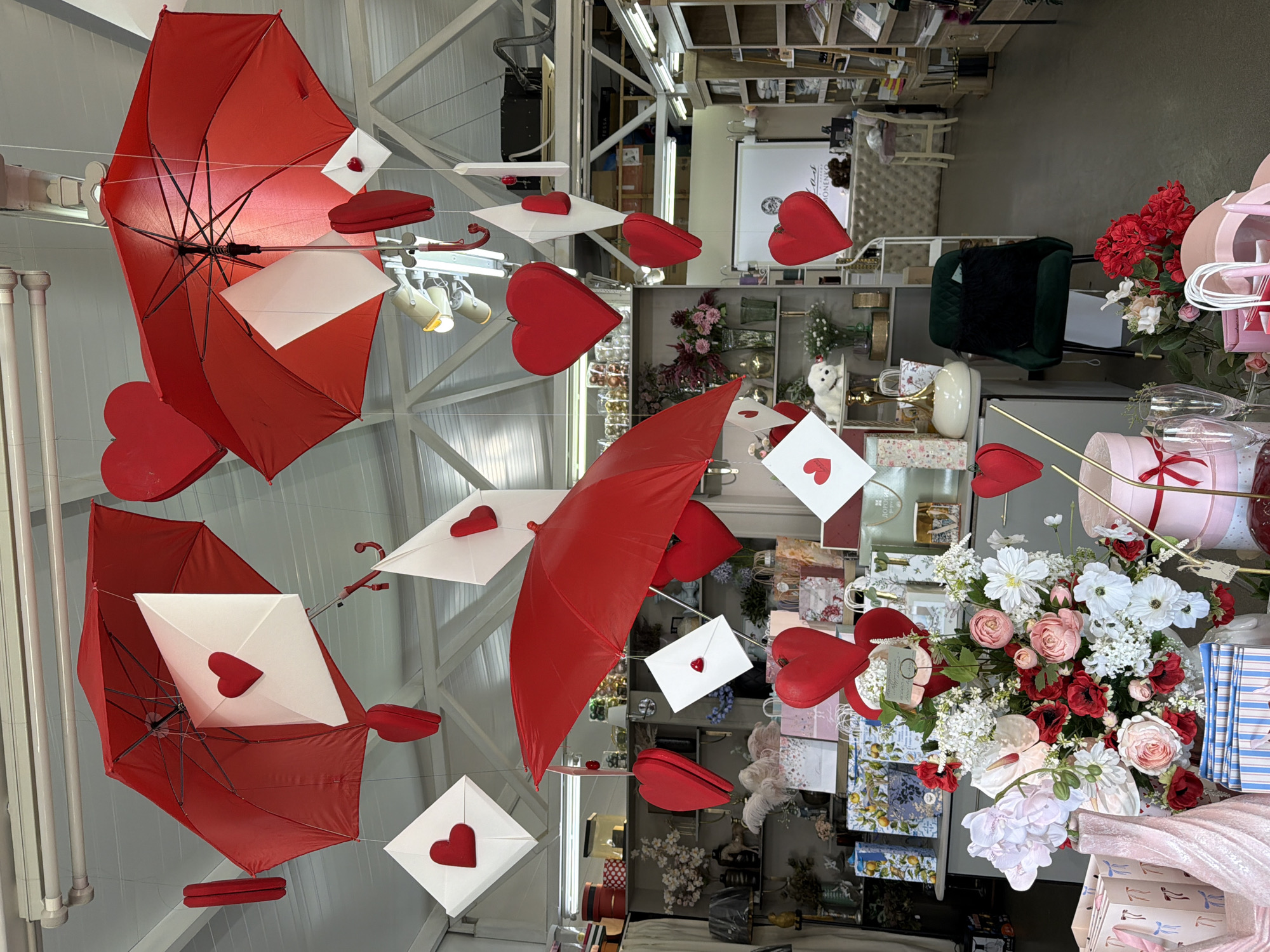 The scene depicts the interior of a retail establishment, identified as being in Votkinsk, Russia, adorned with Valentine's Day themed decorations. Suspended from the ceiling are multiple inverted red umbrellas, intermingled with white square envelopes, each featuring a red heart on its exterior, and numerous additional red heart-shaped decorations. The ceiling structure reveals white trusses and exposed overhead lighting fixtures. In the foreground, a prominent floral arrangement, composed of red, pink, and white flowers, rests on a display table. Surrounding this arrangement and on adjacent surfaces are various retail items, including patterned gift bags, gift boxes (some pink with ribbons), striped notebooks or packages, and glassware. Shelving units line the walls, displaying an assortment of merchandise such as vases, lamps, a white dog figurine, and other decorative objects. In the midground-right, a dark green velvet chair is visible. The background features a light-colored counter, potentially a service or checkout area, with a white sign displaying text and a logo behind it. Additional shelves filled with products extend along the walls further into the establishment. The floor is a consistent dark gray surface. No individuals are present in the image.
