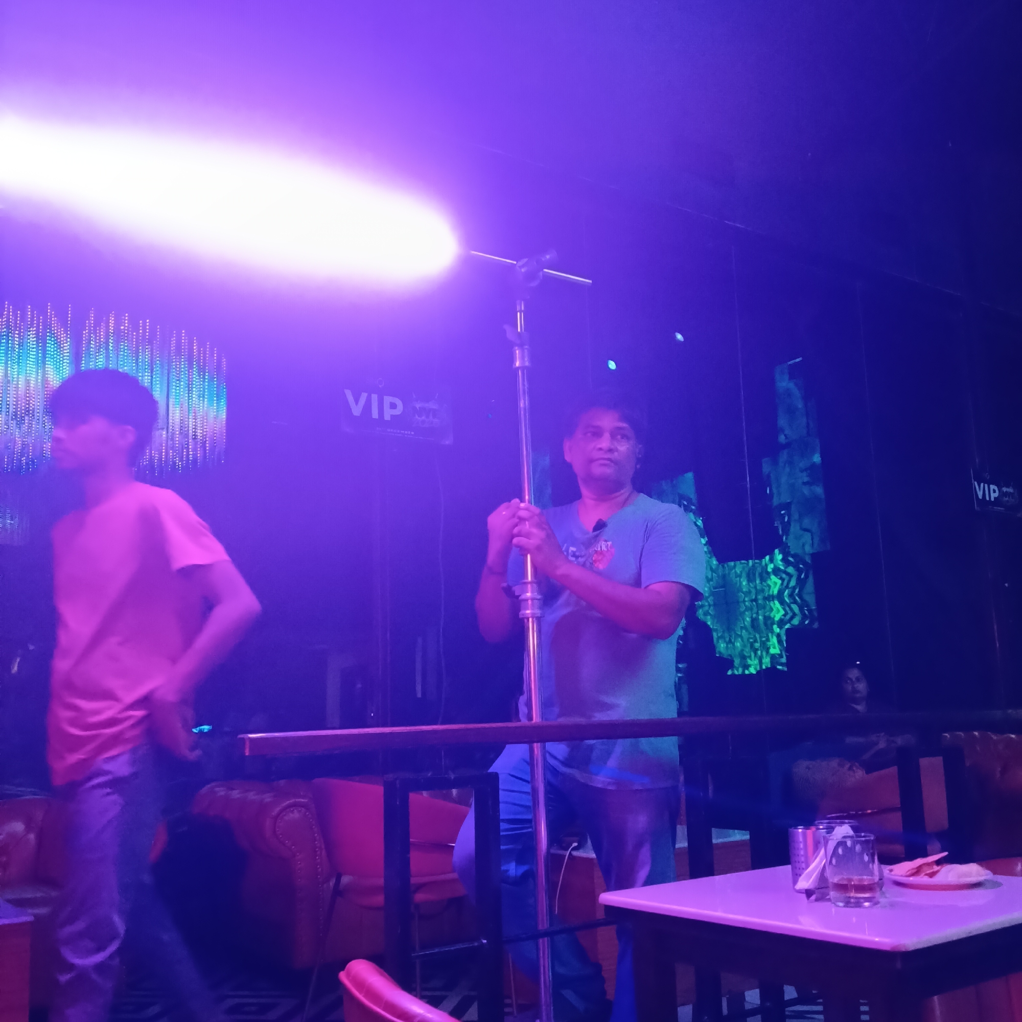 The image shows an indoor scene, likely a bar or nightclub, in Sinquerim, India. The lighting is predominantly blue and purple, creating a club-like atmosphere. In the foreground, a man wearing a blue t-shirt is holding a metal pole that appears to be a light stand or similar equipment. A bright light is emanating from the top of the pole, obscuring details. Another person in a pink t-shirt is walking in the left foreground.
Behind the man with the pole, there's a partial view of a VIP sign and a patterned light display on the wall. In the right foreground, there is a table with glasses and a plate with food on it. Behind the table is a railing that separates the area from a seating area with red leather couches and chairs. A person is seated in the background behind the railing. The room has a black tiled floor.