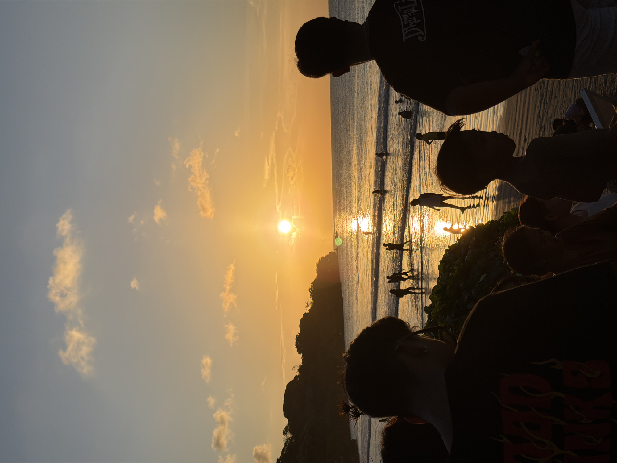 The image depicts a sunset over the ocean in Kuta Selatan, Indonesia. Silhouettes of people are visible in the foreground and on the beach. The sun is setting over the water, creating a bright reflection on the ocean surface. There is a dark land mass between the camera and the sun. There are some small clouds in the sky.