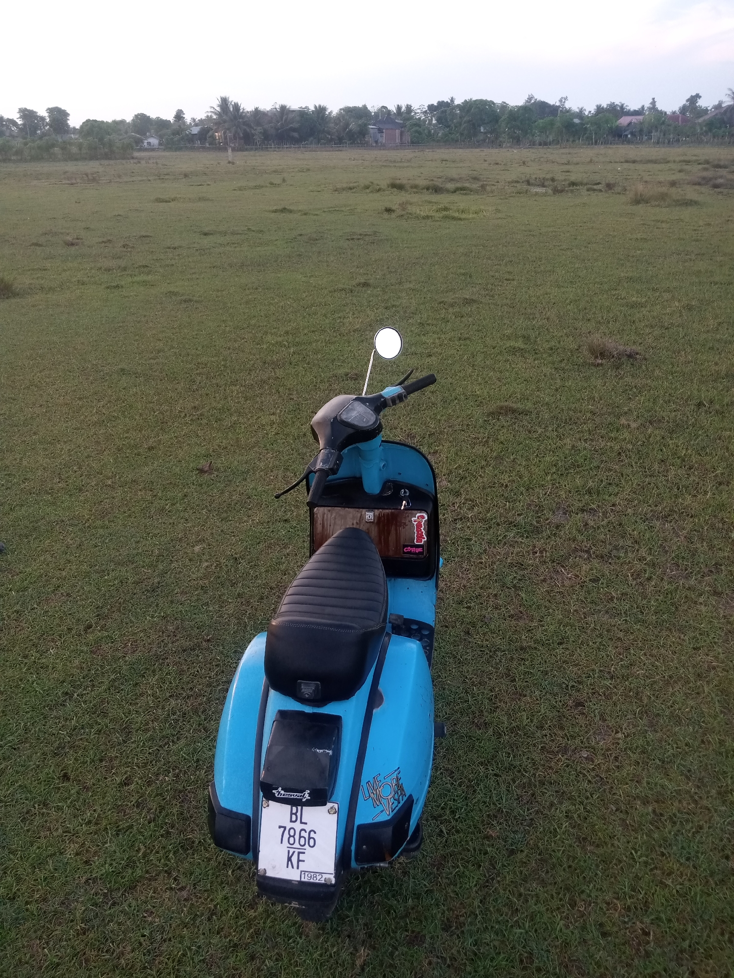A blue scooter is parked on a grassy field in Paya Linteung, Indonesia. The scooter has a black seat and handlebars. A license plate on the back of the scooter reads "BL 7866 KF" and "1982". In the background, there are trees and houses. The sky is overcast.
