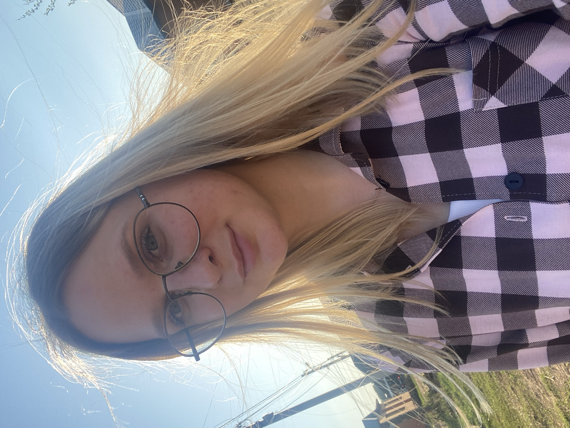 A young woman with blonde hair and round eyeglasses is outdoors in daylight. She is wearing a pink and black buffalo plaid shirt. The background includes a blue sky, power lines, green grass, and a building. Based on the architecture and environment, the location is Chaikovsky, Russia.