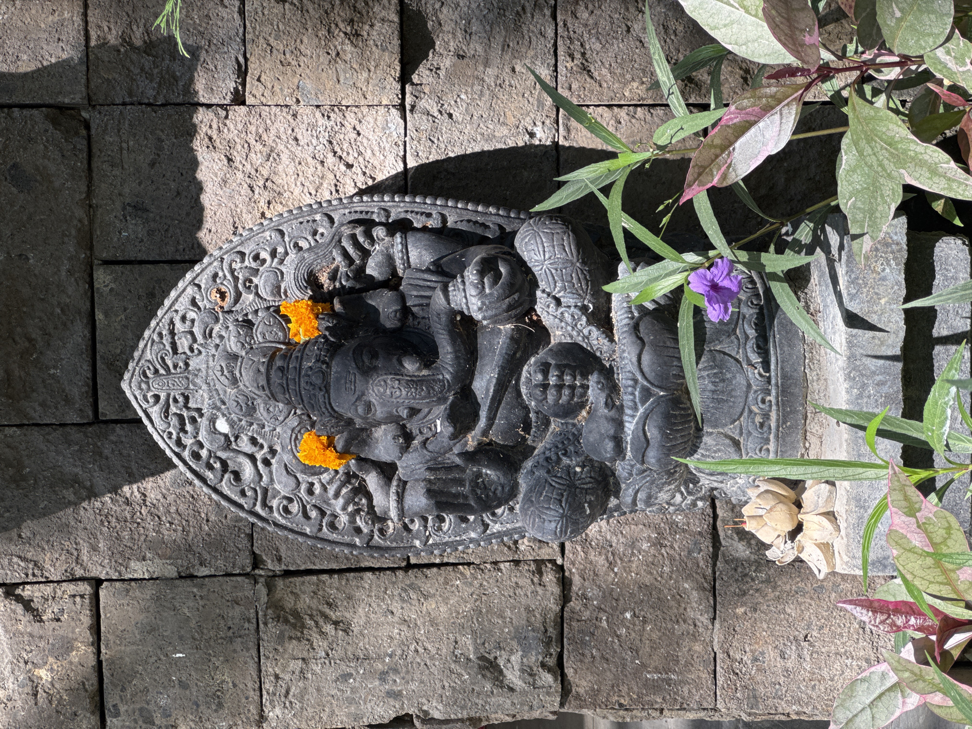 A dark grey stone statue of the Hindu deity Ganesha is centered against a wall constructed from rectangular, rough-hewn dark grey stone blocks. The statue portrays Ganesha in a seated position, featuring an elephant head, four arms holding various attributes, and a crown. Intricate carvings decorate the statue and its arched surround. Two orange marigold flowers are positioned on Ganesha's head. The statue is mounted on a multi-tiered stone pedestal. Direct sunlight illuminates the upper left area of the wall and portions of the statue, creating defined shadows on the lower right wall and the right side of the statue. In the foreground, surrounding the statue's base, are several plants. These include a plant with long green leaves bearing a single purple flower on the left, and another plant displaying variegated green and pink leaves on both the lower left and right. To the immediate left of the statue's base, on a lower stone platform, rests a small, light-colored lotus-shaped object, possibly an incense holder, with a single red incense stick inserted. The scene is located in Kuta Selatan, Indonesia.