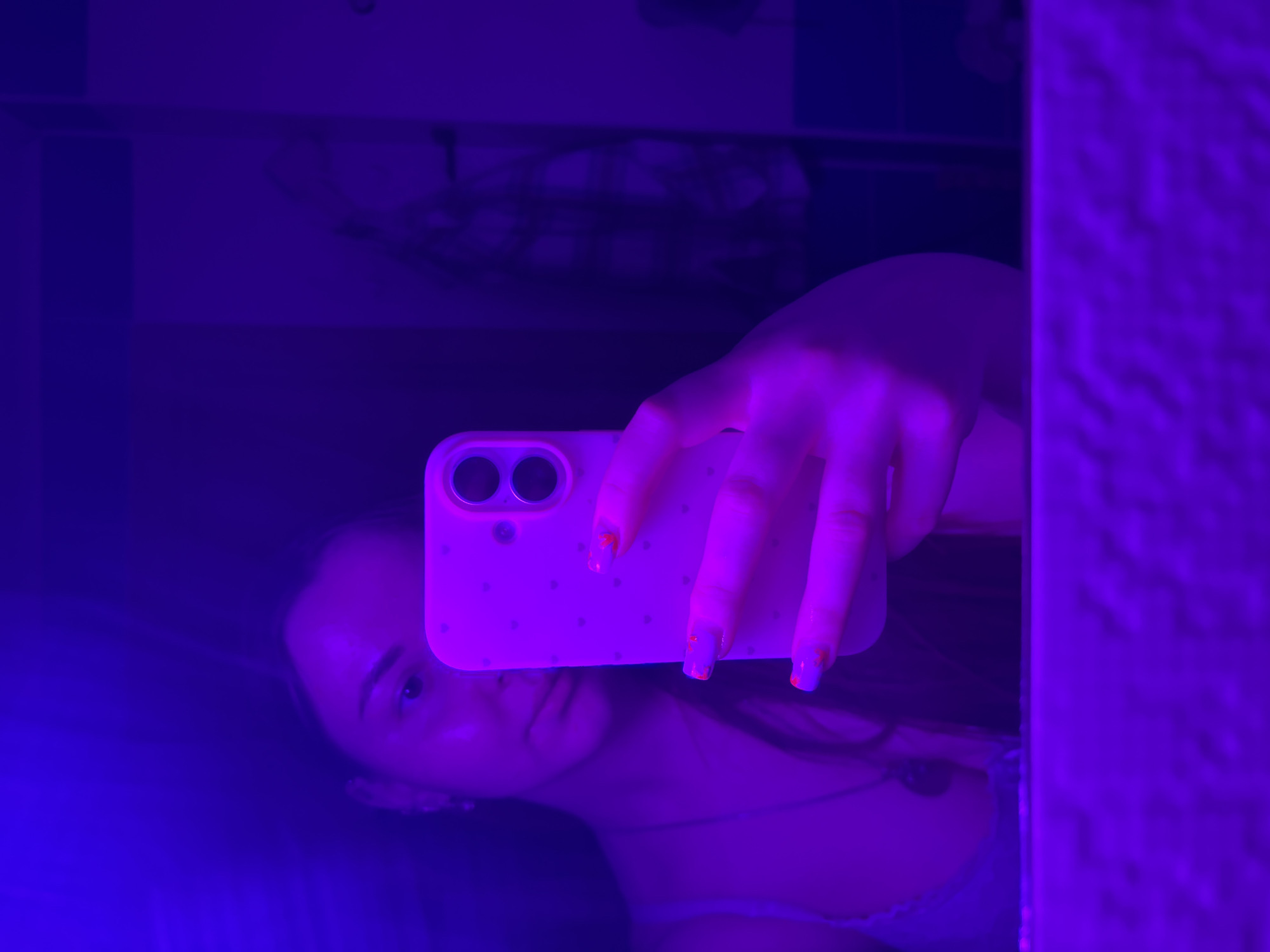 A person, likely a young woman, is captured in a mirror selfie, illuminated by intense purple and blue ambient lighting. Her face, partially obscured by the smartphone she holds, is visible, angled towards the camera's reflection. She has dark hair and appears to be wearing a light-colored top.
Her hand, with fingers displaying manicured nails featuring small orange or red dots, grips a smartphone. The phone has a light-colored case, possibly white or light pink, with two prominent camera lenses on the back and a pattern of small, reflective dots or stars.
The scene is an indoor setting, with blurry, dark reflections in the background suggesting furniture or other room elements. The dominant purple and blue lighting creates a moody, stylized atmosphere, indicating the photo was likely taken in a darkened room, possibly at night. The activity depicted is the act of taking a mirror selfie. No visible text is present in the image. This scene is located in Izhevsk, Russia.