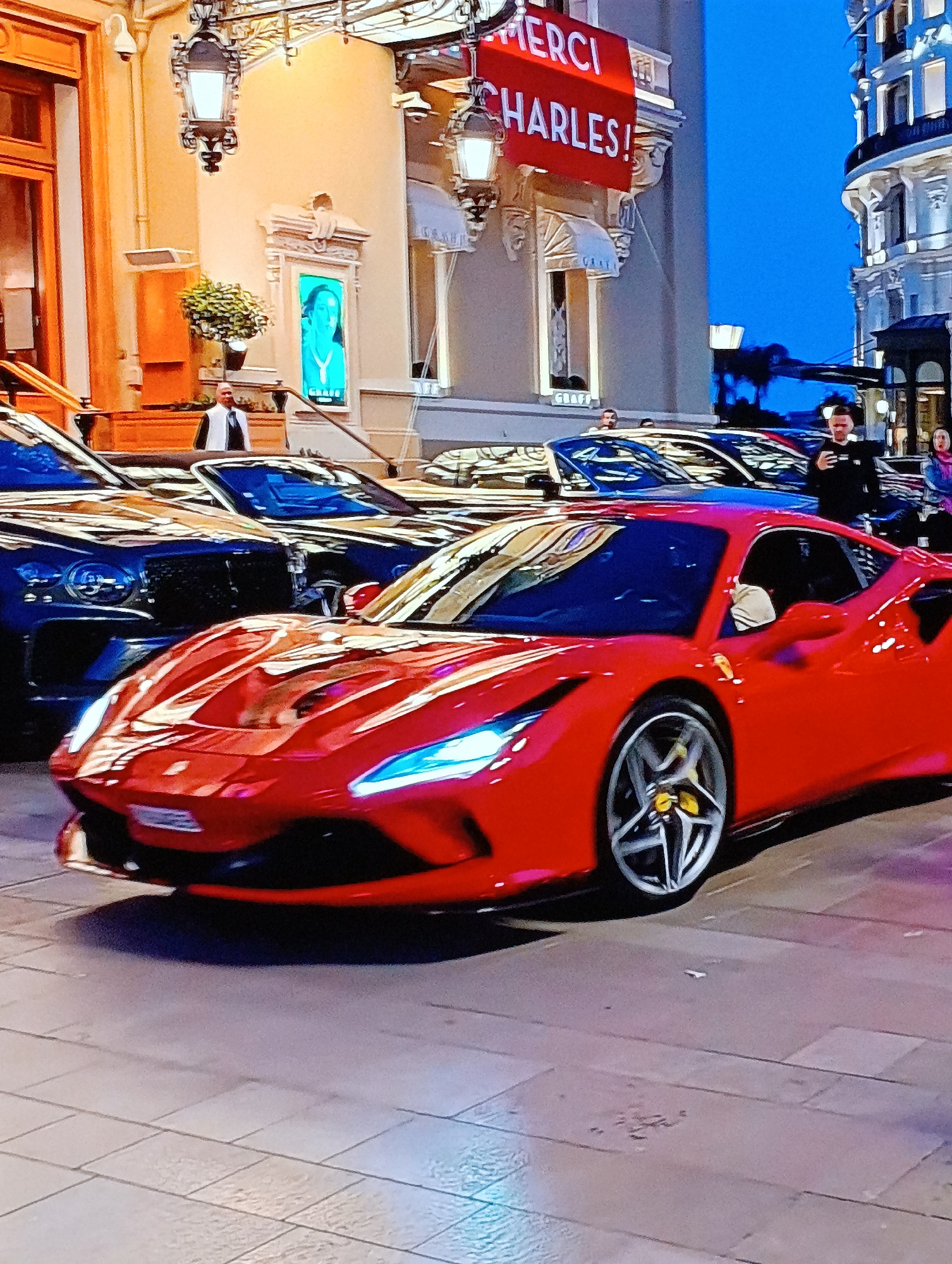 Here is a description of the image:
The image shows a street scene in front of a building. There is a red Ferrari car in the foreground. Behind the Ferrari are other luxury cars, including at least one Bentley. A man in a black jacket is visible to the right of the Ferrari, holding a phone and appearing to take a picture. In the background, a building with ornate architectural details is visible. A red banner with the words "MERCI CHARLES!" hangs from the building. The location is likely Monaco.