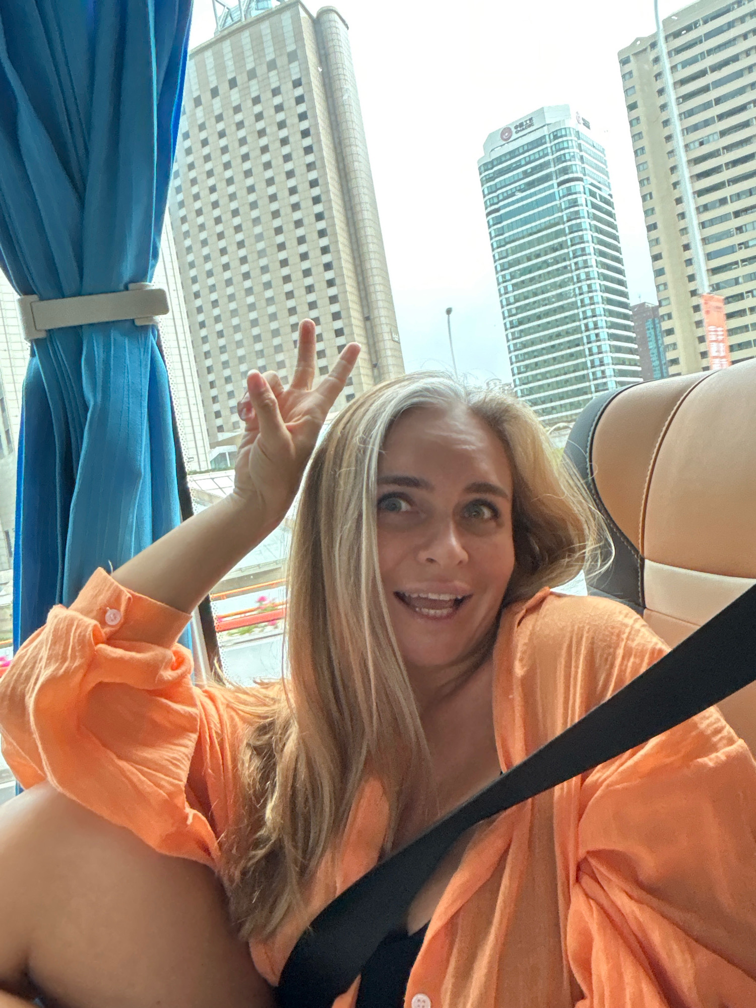 An outdoor, eye-level, medium shot shows a woman in a bright orange, button-up shirt with her right arm raised and fingers extended in a peace sign. She is in the passenger seat of a vehicle, indicated by the seatbelt across her body and the interior of the vehicle visible in the foreground. The woman has blonde hair with streaks of gray, and she is looking directly at the camera with a surprised expression, mouth open. The background is a cityscape with tall buildings, including one with a sign that appears to be in Chinese characters and another that displays the Bank of China logo. The setting is identified as Changning District, China. The overall lighting is bright, suggesting daylight.