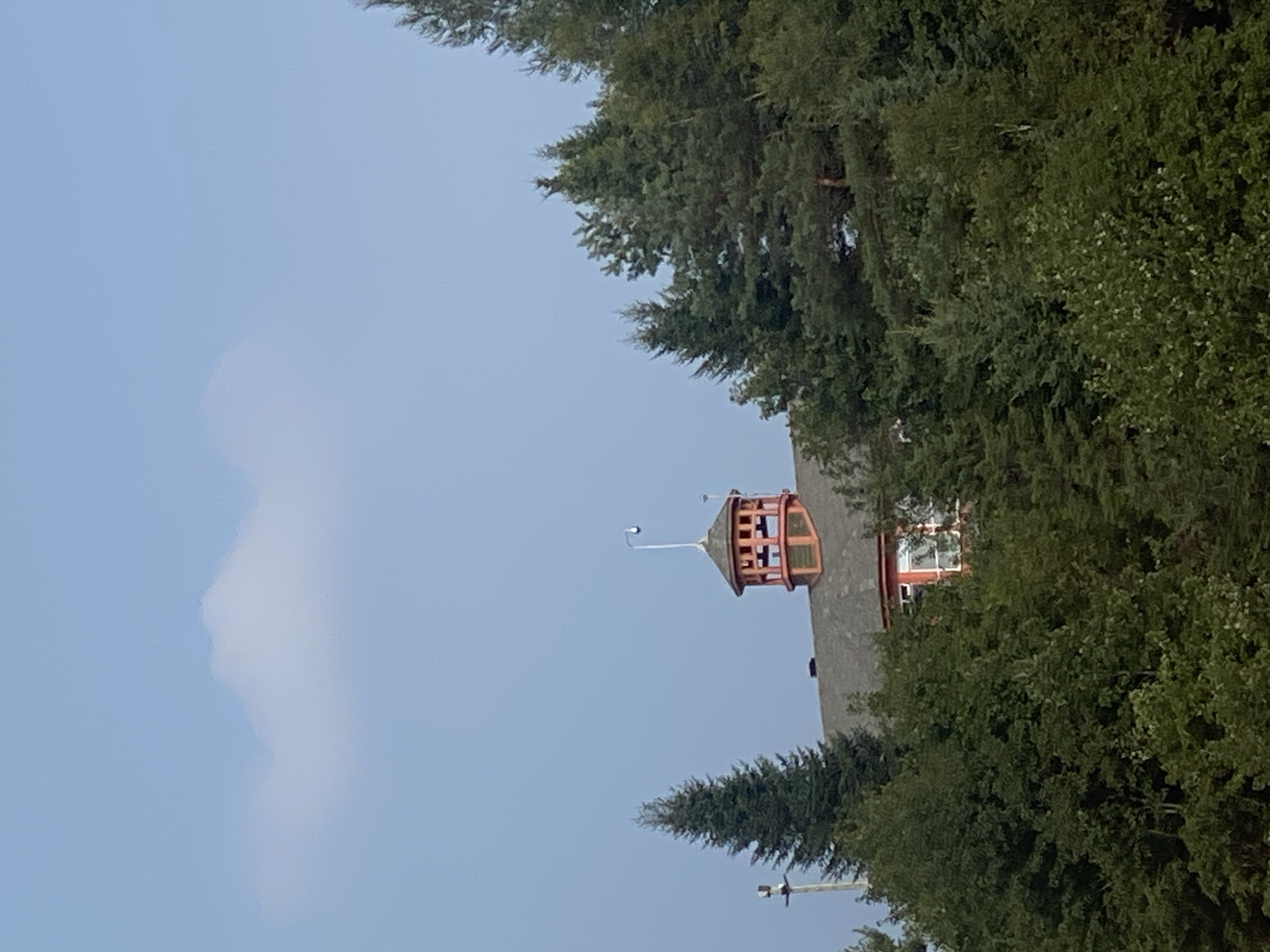 The image shows a building with a distinctive red and green cupola, partially obscured by a dense forest of evergreen and deciduous trees. The building has a grey roof and visible windows, one with white framing. A security camera is mounted on a pole extending from the cupola. The sky above is a pale blue with a single wispy white cloud. The scene appears to be set in Saint-Gabriel-de-Valcartier, Canada, given the prevalence of forested areas typical of the region. No people are visible in the frame.