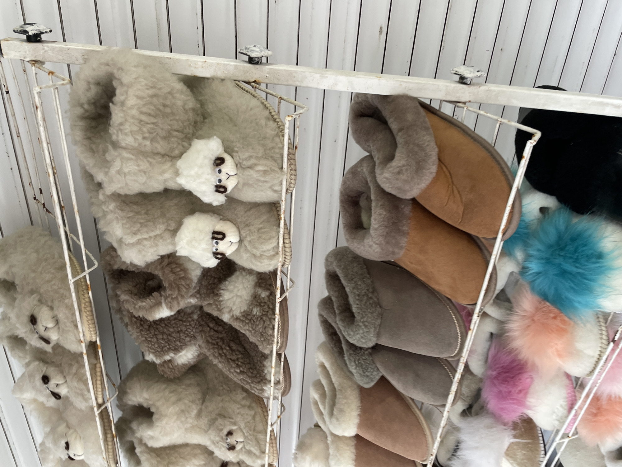 The image shows a retail display of indoor slippers in what appears to be a shop. The slippers are arranged on a white, metal rack with a slightly distressed or aged appearance.
On the left side of the rack are several pairs of fluffy, light-colored slippers with small alpaca or llama-like faces attached to the front. The faces are white with black eyes and stitched details.
The right side of the rack displays a variety of other slipper styles. There are brown and gray slippers with fur-like trim. Additionally, there are slippers with colorful, fluffy pompoms, including shades of blue, pink, and white.
The background consists of vertically aligned white planks.
The location is Olginka, Russia.