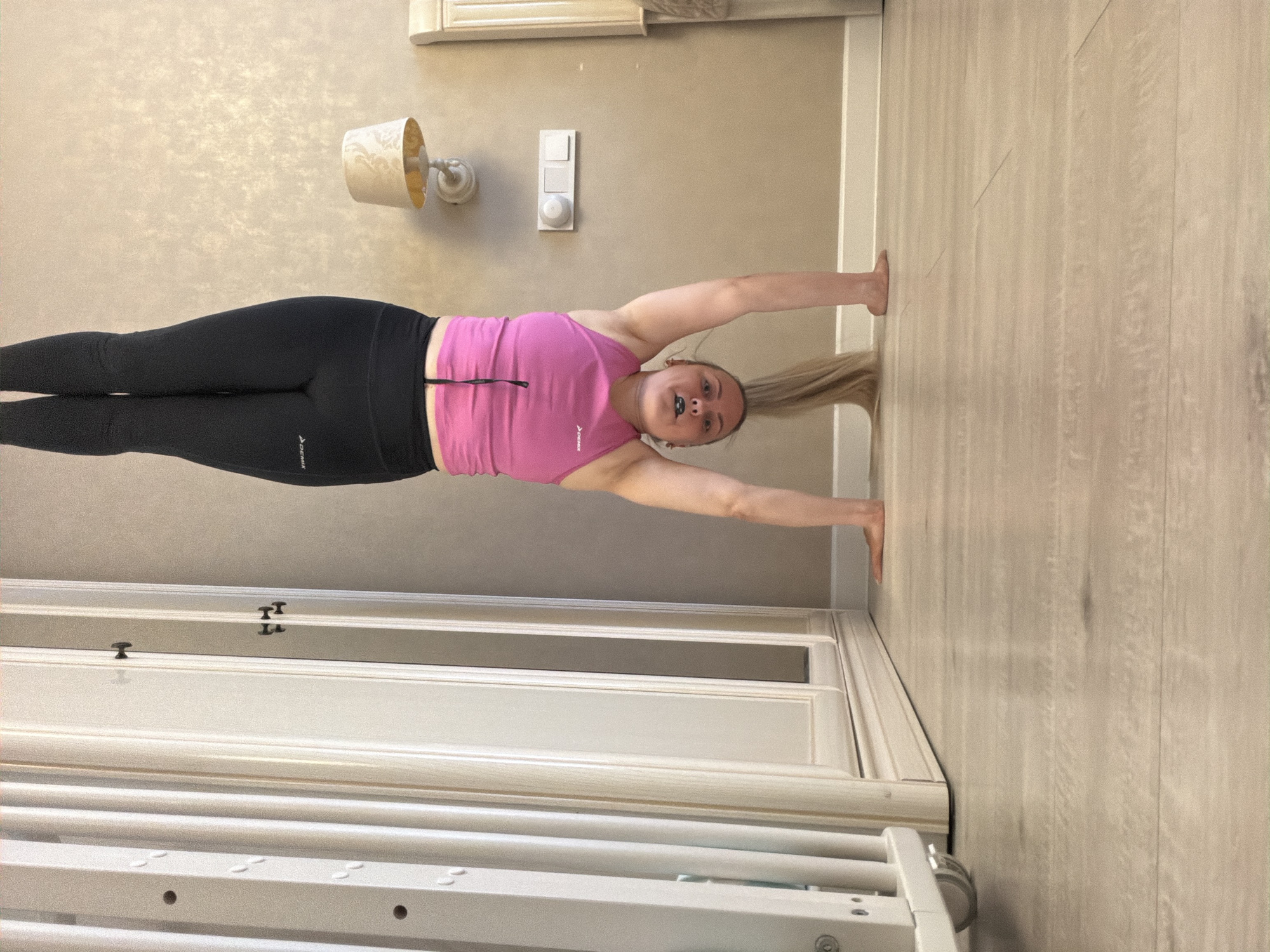 A young adult female is performing an exercise or stretch in an indoor setting, likely a home in Votkinsk, Russia. The image is rotated 90 degrees clockwise, showing the person horizontally across the frame. Her body is straight, stretched between a wall and a wooden floor. Her feet are pressed against a light-colored, textured wall, while her hands are extended behind her head, pressing against a light wooden floor. Her long blonde hair is in a ponytail, extending towards the floor. She is wearing a pink sleeveless athletic top and black full-length athletic leggings. Both the top and leggings feature a white "DEMIX" logo.
Her face is turned slightly towards the camera, revealing a neutral expression. The room has light beige or cream-colored walls with a subtle pattern. A wall-mounted lamp with a decorative shade and a white electrical switch panel are visible on the wall. A white door frame is present to the right. In the bottom foreground, part of a white furniture item, possibly a crib or changing table with railings and wheels, is visible. The lighting appears bright and even, consistent with an indoor environment.