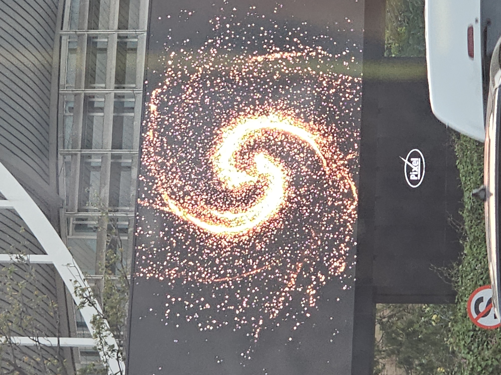 A large digital billboard displays a swirling yellow and orange galaxy-like pattern against a dark background. Below the main display, a smaller oval logo with the word "Pixel" in white lettering is visible. The billboard is situated above a hedge and a street. In the foreground, the tops of two vehicles are visible, one white and one dark. To the left of the vehicles, a circular red and white sign with a black "S" inside a red circle with a diagonal line through it is partially visible. The background shows a building with a metal roof and glass windows, with some trees and branches in front of it. The scene appears to be outdoors in a city setting, possibly Kuta, Indonesia, given the context provided. There are no people clearly visible in the image.