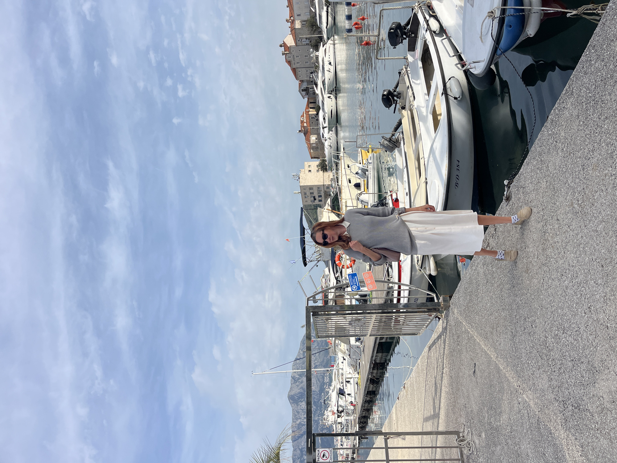 A woman stands on a concrete pier next to docked boats at the marina in Budva, Montenegro. She is wearing sunglasses, a gray sweater, and a white skirt. The sky is partially cloudy and there are mountains in the distance. Buildings are visible on the opposite side of the water.