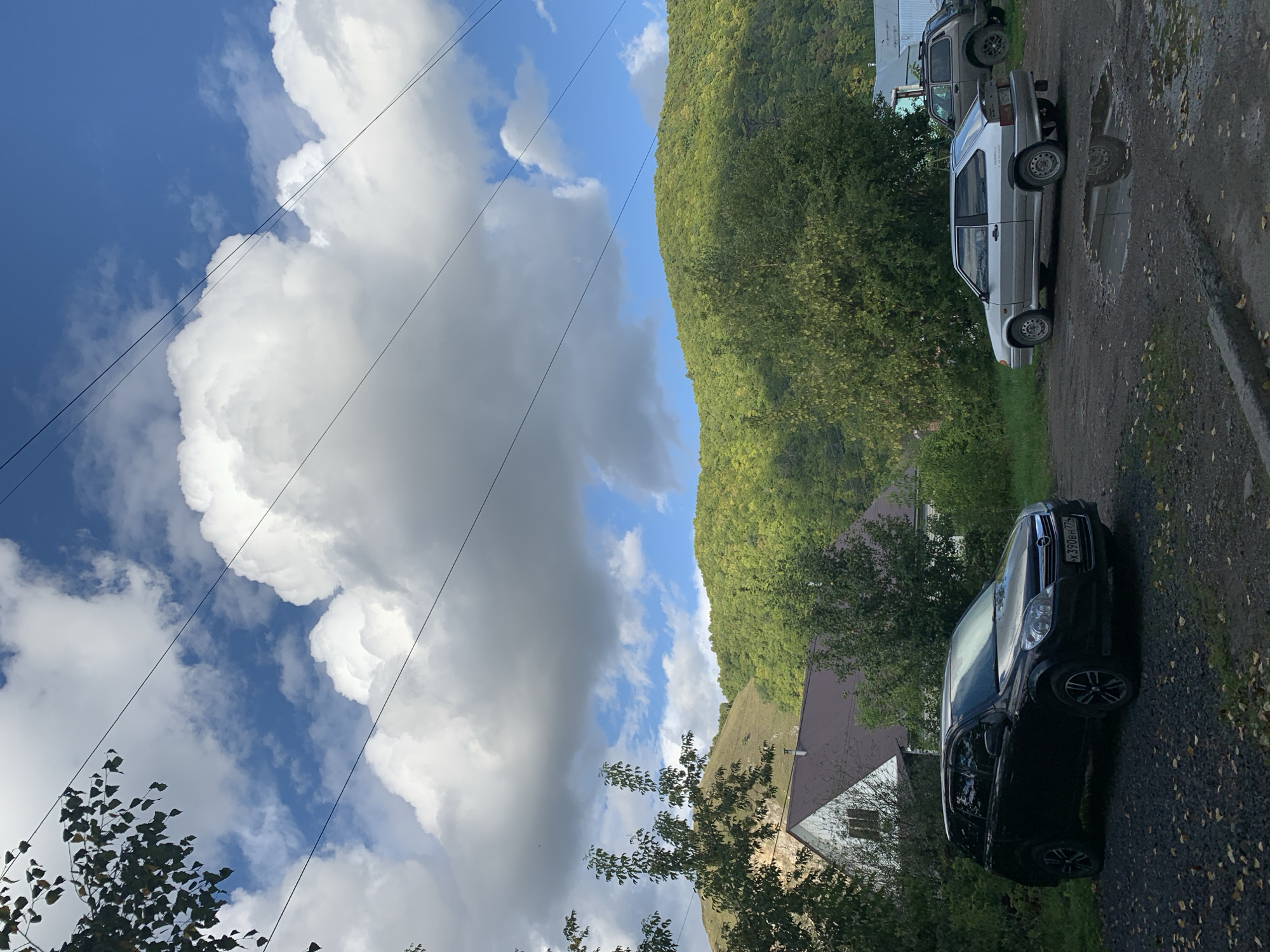 A black Opel Astra station wagon is parked on a gravel lot in front of a white house with a dark brown roof. To the right of the Opel, a silver Lada Samara hatchback is parked, with a brown SUV partially visible behind it. The scene is set against a backdrop of lush green hills and a bright blue sky with dramatic white clouds and overhead power lines. The location is the city of Asha, Russia.