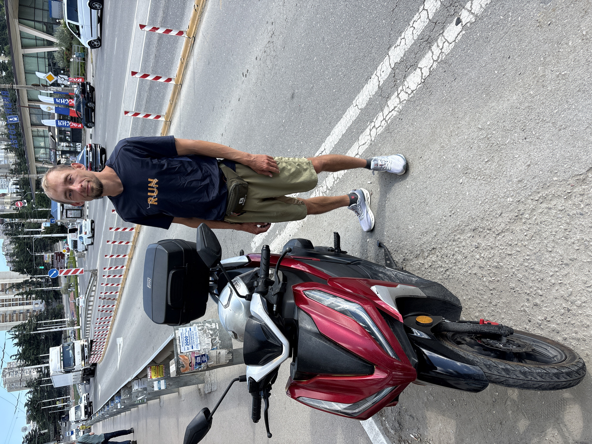 A man stands next to a red and black scooter on a paved road, with buildings and trees in the background. The man is wearing a dark blue t-shirt with "RUN" written in orange, khaki shorts, a green fanny pack, and grey athletic shoes. The scooter is a modern model with a black storage box on the back. The scene appears to be in a city, with a road, traffic, and buildings. There are red and white striped bollards along the side of the road, and a street sign with Cyrillic text is visible in the background. The sky is overcast.