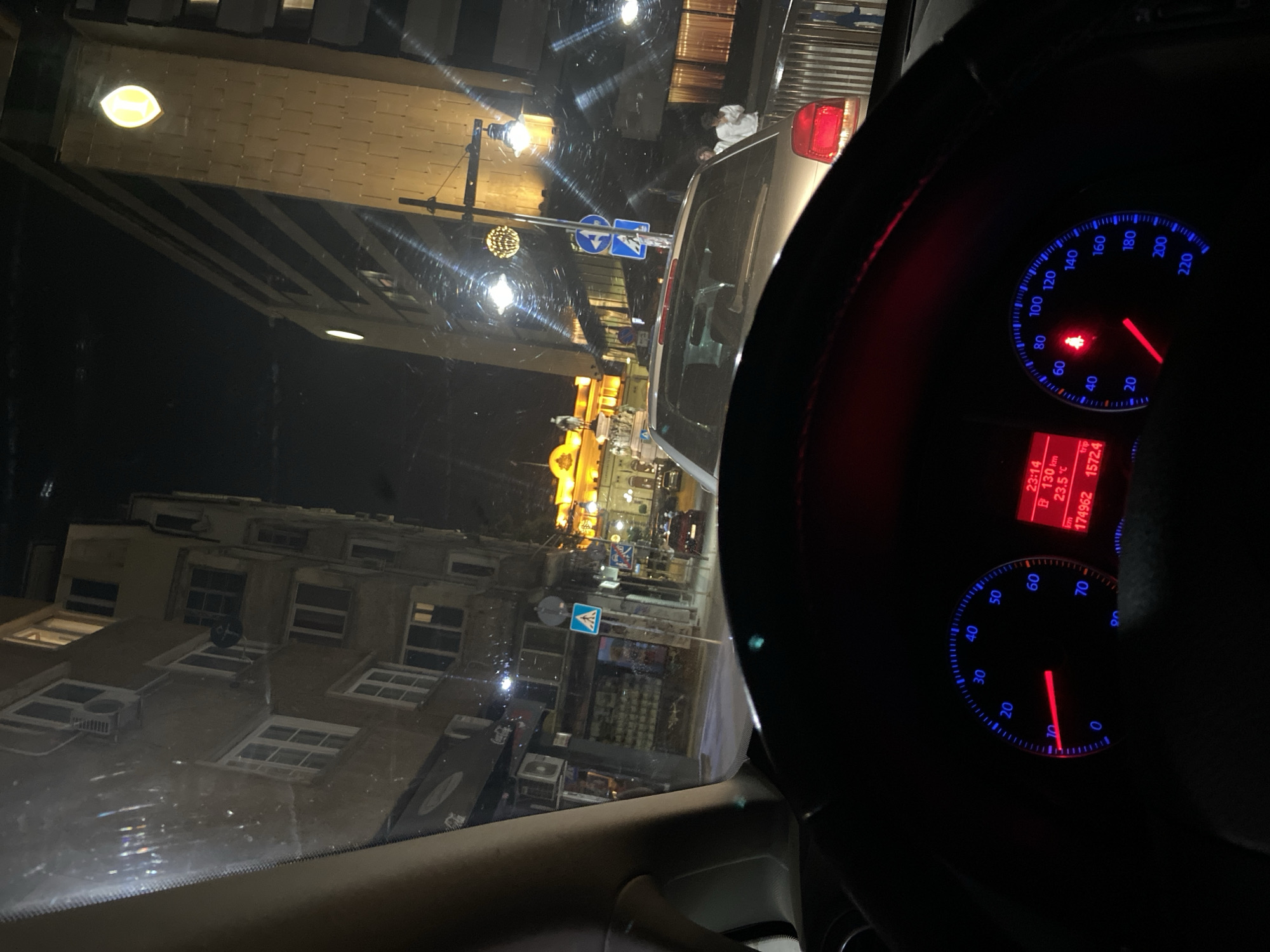 The image presents a nighttime street scene in Sofia, Bulgaria, viewed from inside a vehicle. The car appears to be stationary or moving very slowly, indicated by the speedometer showing approximately 5 km/h and a low tachometer reading. The digital display on the dashboard indicates a time of 23:14 (11:14 PM), an ambient temperature of 23.5 °C, an odometer reading of 174962 km, and a trip meter of 1572.4 km. A red seatbelt warning light is illuminated.
The location is Narodno Sabranie Square. To the right, a prominent building with the illuminated "I" logo of the InterContinental Hotel Sofia is visible, characterized by its golden-toned facade. Further down the street, directly ahead, the brightly lit yellow facade of the National Assembly (Parliament) building is visible, with the equestrian Tsar Liberator Monument in front of it.
In the midground, a light-colored car is positioned directly ahead of the viewer's vehicle, its red taillights illuminated. Two individuals are standing on the sidewalk to the right of this car, wearing light-colored tops. Several streetlights illuminate the area. Various street signs are present, including a blue square pedestrian crossing sign on the left and a blue circular mandatory right turn sign with a directional arrow below it on the right.
On the left side of the street, multi-story buildings with numerous windows and ground-level storefronts are visible. One storefront displays a "Coca-Cola" logo. An air conditioning unit and a small satellite dish are mounted on one of these buildings. The sky is dark, consistent with the nighttime setting. The car's windshield shows reflections and some minor splatters or dirt.