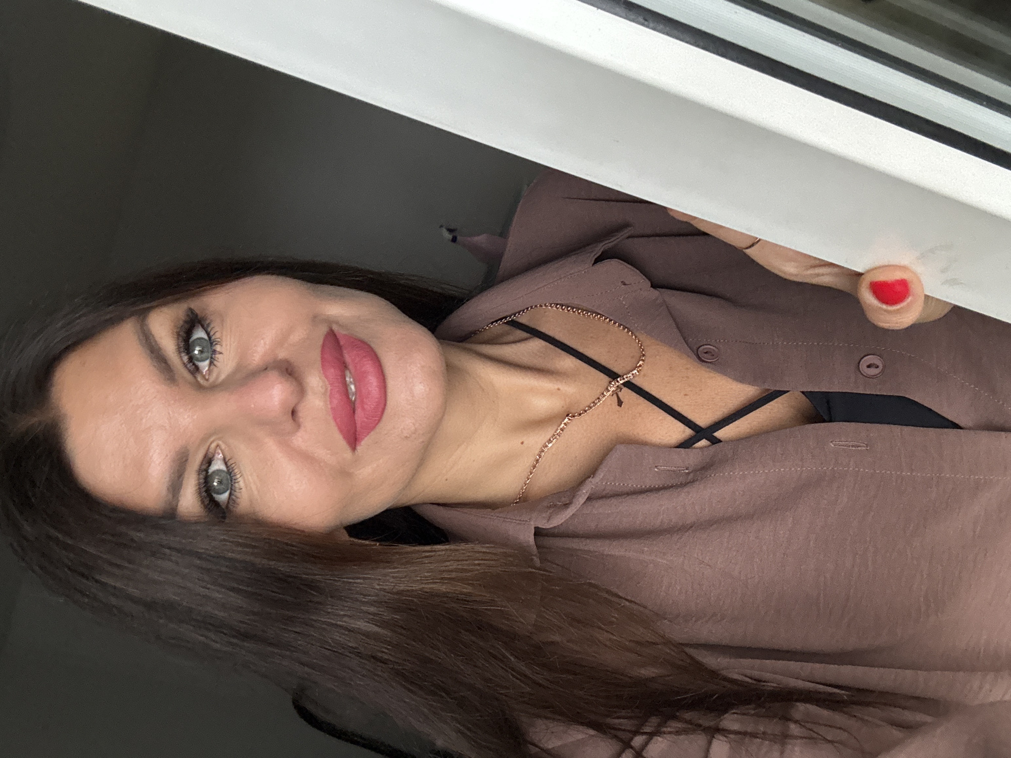 A close-up shot shows a woman with long brown hair and blue eyes looking into the camera. She is wearing a brown blouse with a black bra underneath, and a gold necklace. Her lips are painted a bright pink. She is holding onto a white object, possibly a door frame, with her right hand, which has red nail polish on her nails. The background is dark and out of focus. There are no discernible elements that indicate this photo was taken in Chaikovsky, Russia.