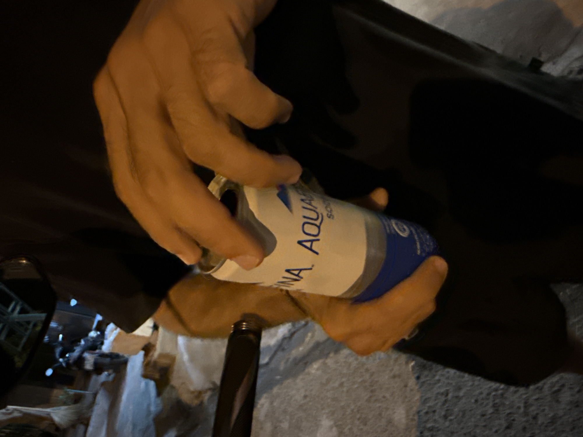 A person, likely male based on hand size and hair texture, is holding a can of what appears to be Aquafina sparkling water. The can is mostly white with blue lettering, including "NA AQu". The person's hands are visible, one gripping the can from the side and the other near the top, suggesting they are either opening or holding it. The background is dark and appears to be outdoors at night, with a blurred motorcycle visible in the distance. The ground is rough and grey, consistent with a street or paved area. The lighting is warm and orange-toned, possibly from artificial sources. The image is focused on the hands and the can, with the background out of focus. The location is specified as the city of Nha Trang Ward, Vietnam.