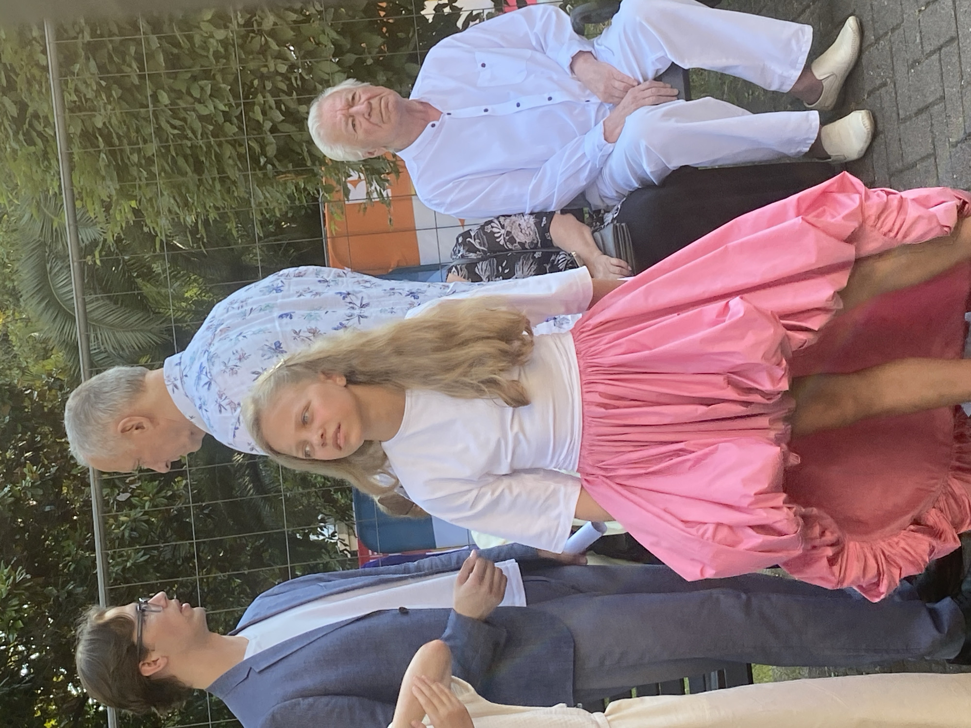 The image depicts an outdoor scene with four individuals, suggesting a gathering or event. In the foreground, a young girl with long blonde hair wears a white t-shirt and a voluminous, bright pink skirt. To her left, a young man in a blue blazer over a white shirt and gray pants faces away from the camera, engaged in conversation with an older man. The older man, also facing away, wears a white shirt with a floral pattern. To the right of the girl and further back, an older man with white hair is seated on a bench, wearing a white collared shirt and white pants. He is looking towards the left of the frame. The background features a metal fence and lush green foliage, indicative of an outdoor setting, possibly a park or garden. The ground appears to be paved with interlocking bricks. The lighting suggests it is daytime, with sunlight casting shadows. The overall atmosphere appears casual yet organized. The location is identified as Sochi, Russia, implying the context of the event could be a festival, performance, or public gathering in the city.