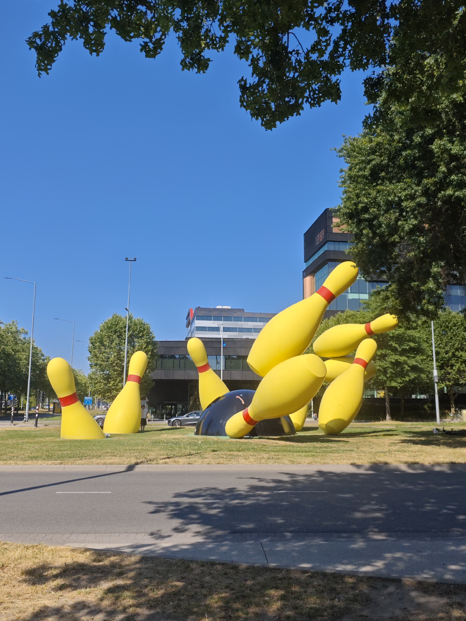 This image depicts the "Bowling Pins" art installation located on the John F. Kennedylaan in Eindhoven, Netherlands. The installation consists of several oversized, bright yellow bowling pins with red bands, scattered and tilted as if knocked over. One black bowling ball is partially visible amongst the pins. The scene is set outdoors on a sunny day with a clear blue sky. In the background, modern glass-fronted buildings are visible, characteristic of the city's architecture. Several trees with lush green foliage frame the top and right side of the image. A paved road runs across the foreground, with dry grass and shadows of leaves on its surface. A few cars are parked or driving on the road in the middle ground, and a lone person stands near the art installation. The overall environment suggests an urban setting with a touch of whimsical public art.