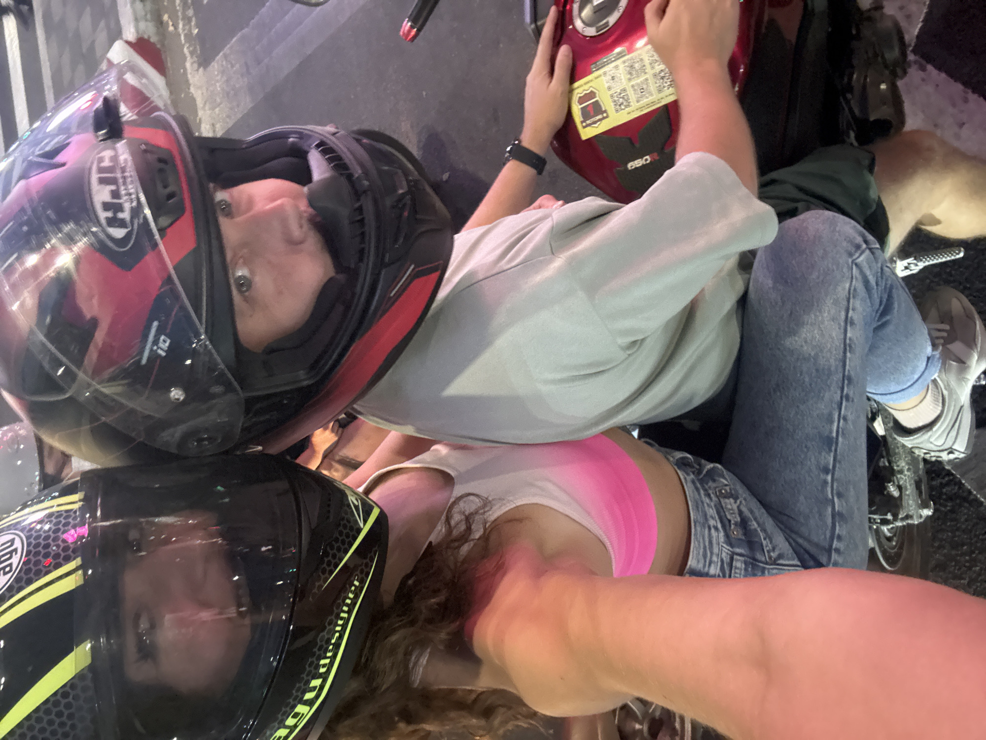The image is a high-angle selfie capturing two individuals on a red motorcycle, likely in Đà Nẵng, Vietnam, during the evening or night. The perspective looks down onto the two subjects, both wearing full-face motorcycle helmets.
The person at the top of the frame, appearing to be male, wears a red and black HJC i10 helmet with the visor slightly open, revealing his face with a neutral expression. He is dressed in a light gray t-shirt and blue jeans, with a dark wristwatch on his left wrist. His right hand rests on the red motorcycle tank, which is adorned with a yellow sticker displaying a "VICTORY" logo, the model number "650R," and two QR codes.
Below him, positioned as if riding pillion, is a second person, likely female, wearing a black and yellow AGV designer helmet. Her face is partially obscured by reflections on the visor. Long, wavy brown hair is visible around her neck and shoulders. She wears a white tank top over a bright pink sports bra or top, paired with denim shorts. Her arm is extended, indicating she is holding the camera for the selfie.
The scene takes place outdoors on a dark, paved surface, suggesting a street or parking area. The artificial lighting creates reflections on the helmet visors. The overall impression is of two people pausing during or before a motorcycle ride, capturing a casual moment together.