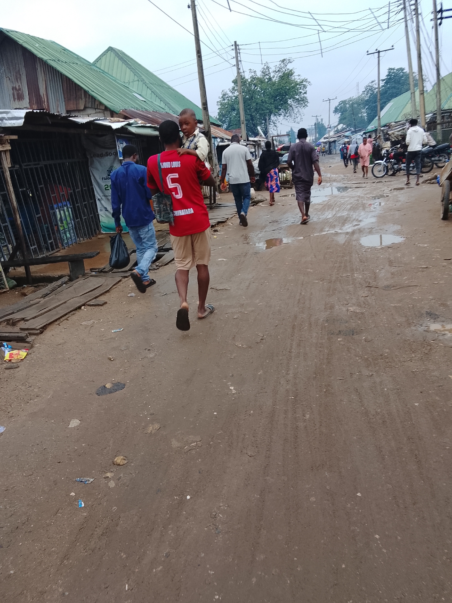In Abuja, Nigeria, a young Black male, perceived to be a teenager, carries a young Black male child on his shoulders, walking away from the camera on a dirt road. The older male wears a red t-shirt with white lettering that reads "LIOMU LOUIS" and a white number "5" on the back, with white text below it that reads "CHIKATARA." He also wears tan shorts and dark flip-flops. The child on his shoulders wears a white and blue checkered shirt and a dark belt.
The scene depicts a moderately populated street, likely in a less developed area of Abuja. Other individuals are visible in the background, walking in various directions. To the left of the frame are several makeshift structures with corrugated metal roofs, some appearing to be shops or stalls. One stall displays a banner with text, partially obscured. Further down the street, more buildings and power lines are visible. The road is unpaved and shows signs of recent rain, with puddles of water and mud. Discarded items, including plastic bags and wrappers, are scattered on the ground. In the distance, parked motorcycles suggest transportation in the area. The overall atmosphere is overcast, with a muted color palette dominated by earth tones and the green of the rooftops.