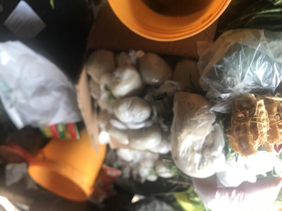 A close-up view of a pile of white, round objects, likely food, wrapped in plastic bags. There is another large plastic bag containing some kind of brown product. The objects are contained within a cardboard box. To the left of the box, there are two yellow buckets partially visible. The objects are piled high, and there are various other plastic bags containing assorted items. The scene appears to be inside a market or shop. The location is the city of Umudike, Nigeria.