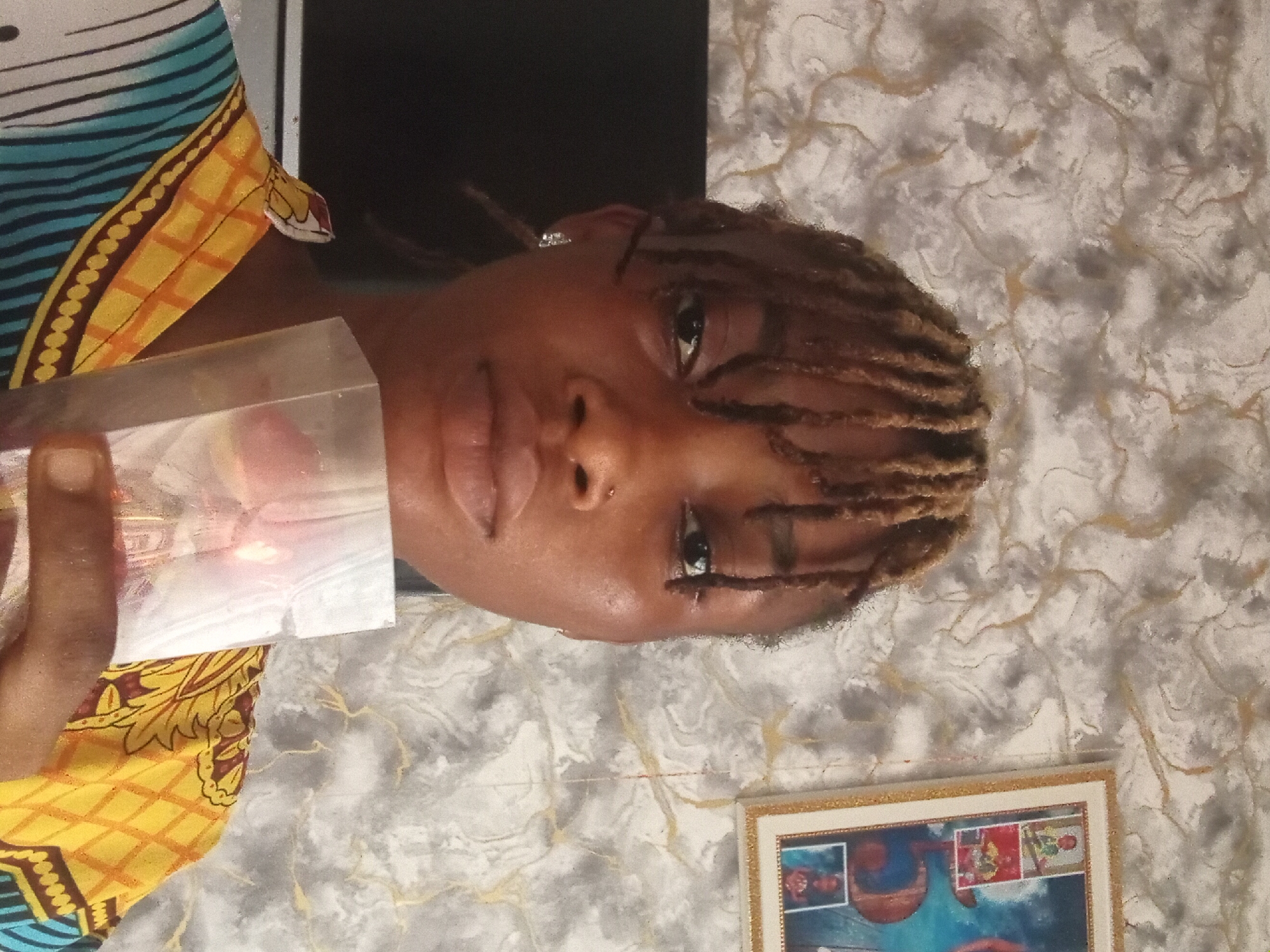 A woman with brown dreadlocks is holding a clear block with a blurred red object inside. She has a nose piercing and is wearing an earring. She's wearing a patterned top with yellow, blue, and brown colors. Behind her is a wall with a marbled grey and white pattern. A framed picture is visible on the wall, below her face. She's indoors. Location, Shagamu, Nigeria is inferred.
