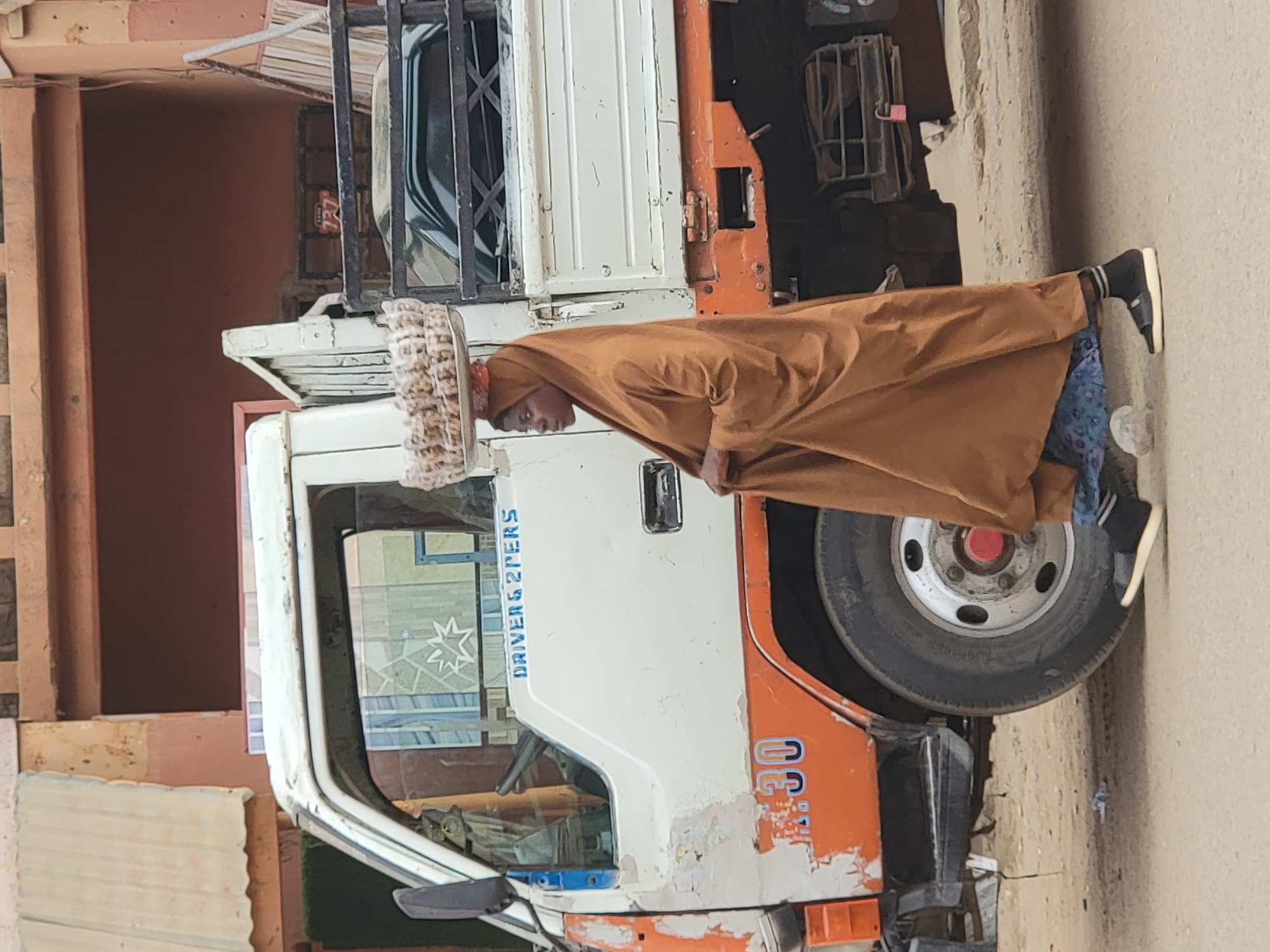 An outdoor image, originally rotated 90 degrees counter-clockwise, depicts a person standing beside a parked truck under natural daylight in Maiburiji, Nigeria.
The central figure is a person, positioned on the right side, facing slightly right and looking downwards. They are clad in a long, light brown garment, likely a traditional robe or jalamia, and a distinctive, light-colored, patterned head covering or hat. Their footwear consists of black shoes with white soles and striped socks.
To the left of the person is a utility or cargo truck, an older model with a white cabin and an orange lower section, showing signs of wear and peeling paint. Visible on the side of the truck's cabin, above the door, is the blue text "DRIVER & 2 PERS". Below this, on the orange bodywork, the faded number "N-30" can be seen. A large tire is prominent on the truck's right side. The reflection in the truck's windshield subtly reveals the eyes of a cat.
In the background, behind the truck's cargo section, part of a reddish-brown building facade is visible, with the numbers "237" faintly discernable on a wall. The ground appears to be a dusty or unpaved surface, and a small white, possibly plastic, object lies near the person's feet. The scene captures a quiet, everyday moment.