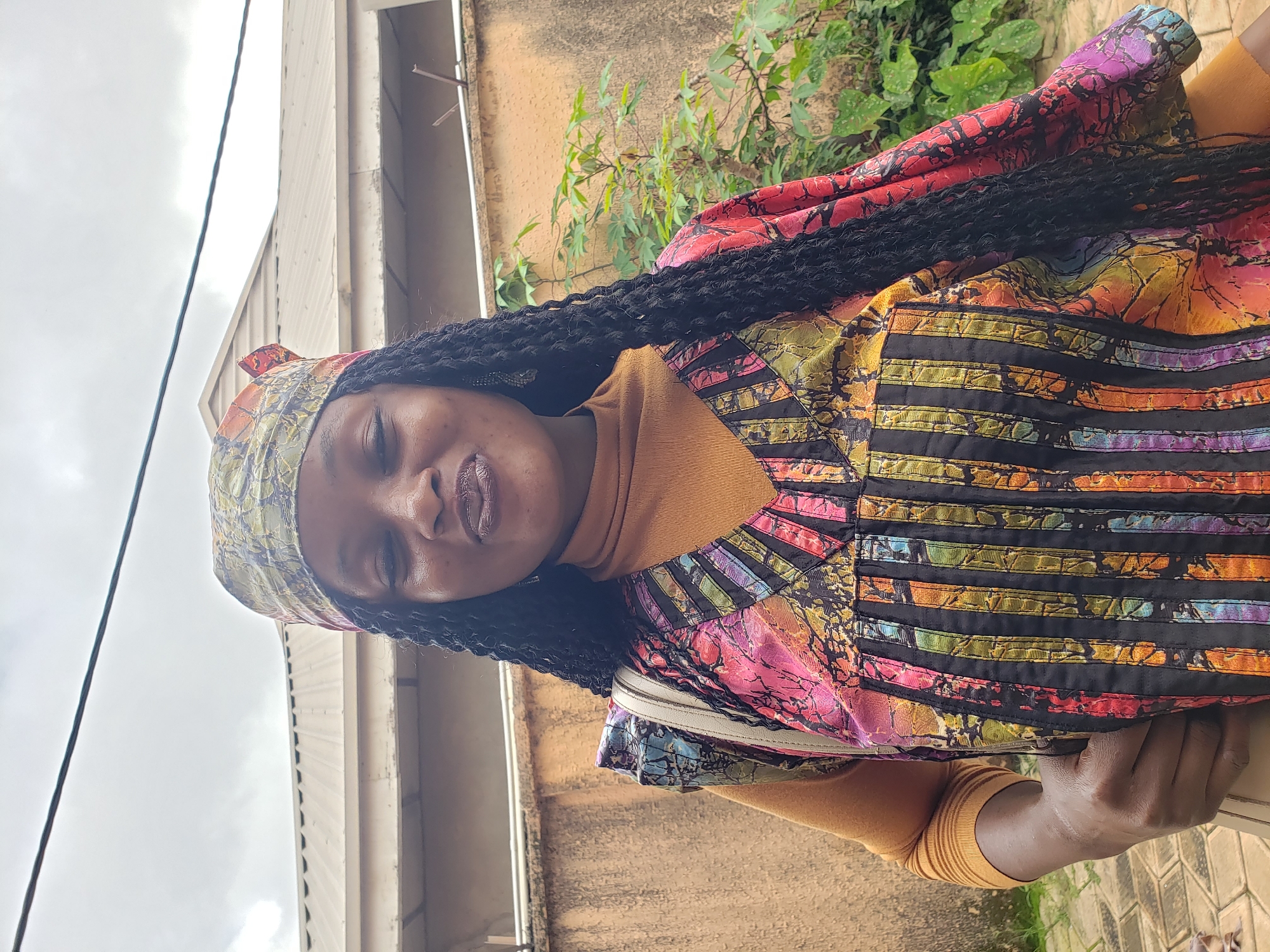 In Sabon Tasha, Nigeria, a woman is captured in a waist-up portrait outdoors. She is wearing a colorful, patterned v-neck top over a mustard-colored turtleneck and a matching headscarf. Her eyes are closed, and she has a slight smile on her lips. Long, black braided hair cascades down her shoulders. Behind her, a muted orange wall forms the backdrop, with a glimpse of a building's corrugated metal roof and some greenery to the right. The lighting suggests an overcast day.