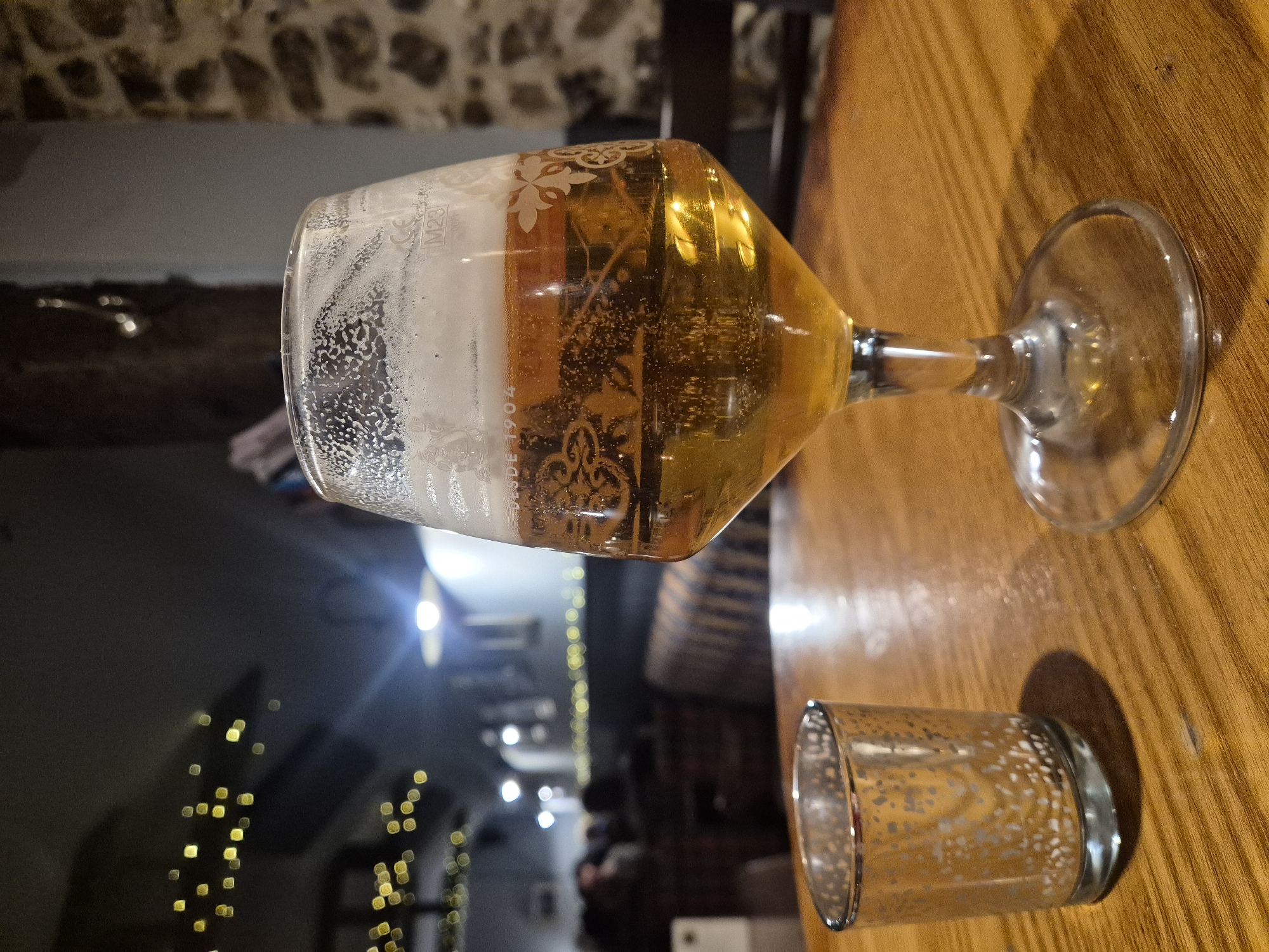 The image depicts a partially consumed pint of beer in a glass, resting on a wooden table. Next to it is an empty glass. In the background, out of focus, is a room decorated with fairy lights. The photo appears to have been taken indoors, possibly in a pub or restaurant. Considering the West Suffolk, United Kingdom constraint, it could be at any drinking establishment in that geographical area.