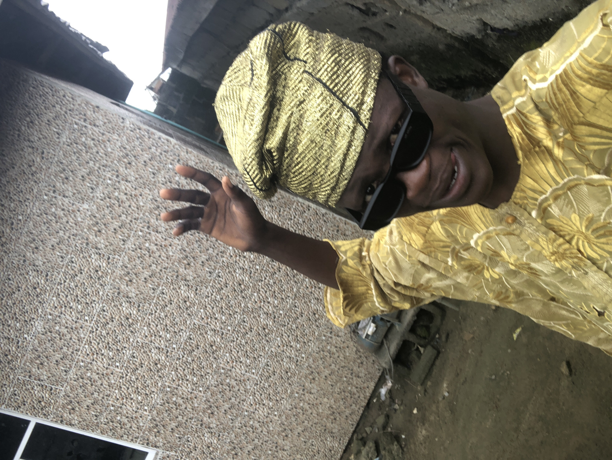 A dark-skinned person wearing a gold brocade cap and sunglasses poses for a selfie outdoors. The person is also wearing a matching gold brocade tunic with ruffled cuffs. Their left hand is raised and open, possibly waving. The background consists of a textured building wall with small, pebble-like stones, a dirt ground with debris, and parts of other structures, including a corrugated metal roof. The overall setting appears to be an urban environment in Ado, Nigeria.