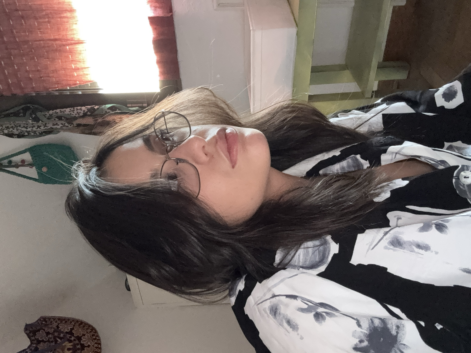 The image shows a young woman with dark hair, wearing glasses and a black and white floral print shirt. She is looking to the right, and her face is partially illuminated by sunlight coming from a window. In the background, there is a green table with a white container on it, a window with red curtains, and some decorative items hanging on the wall. The overall scene suggests an indoor setting, possibly a bedroom or living room. There is no indication of the location being in Las Rozas de Madrid, Spain.