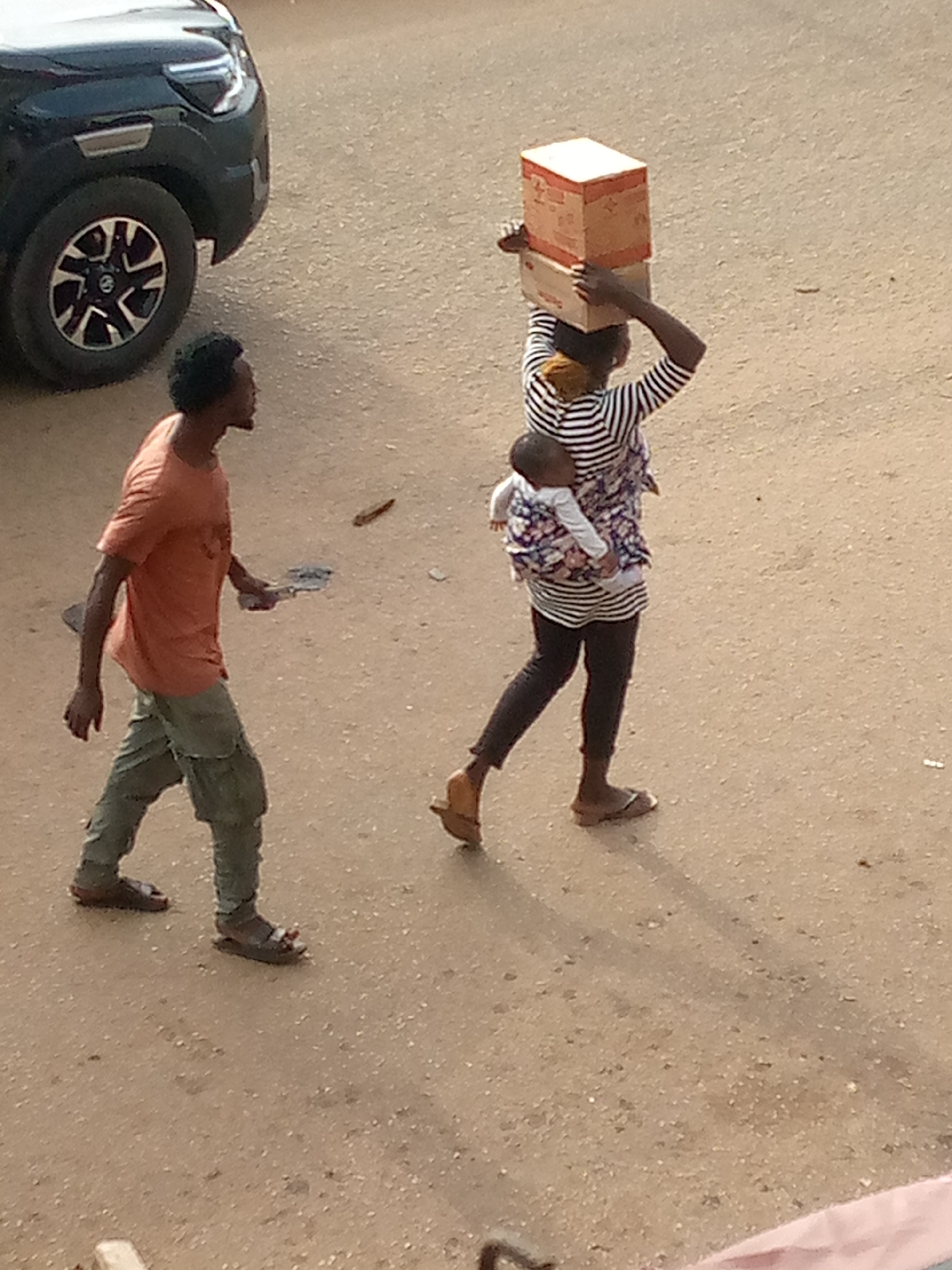 The image shows a man and a woman with a baby walking on a paved surface, likely a street, in Bukuru, Nigeria. The woman is carrying two boxes on her head and has a baby strapped to her back. She wears a striped shirt, dark leggings, and sandals. The man wears an orange t-shirt, green cargo pants, and sandals, and he is carrying a small object in his right hand. A dark-colored vehicle is visible in the upper left corner of the image. The environment appears to be a street scene, with a light brown or tan paved surface, possibly in a residential or commercial area.