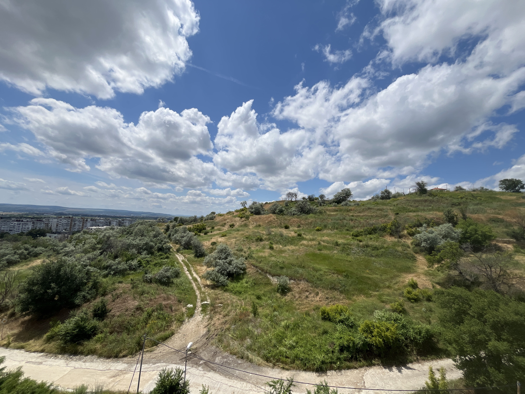 The image shows a hilly, green landscape with scattered trees and a dirt path winding up the slope. In the distance, a city with apartment buildings can be seen. The sky is blue with fluffy white clouds. The scene appears to be on a sunny day. This view is from the area of Varna, Bulgaria, specifically from the southern part of the city, near the Asparuhov most (Asparuhov Bridge), looking inland. The foreground features a paved area, possibly a road or walkway, with some power lines visible. The vegetation is predominantly grass and bushes, with some larger trees scattered across the hills. The overall impression is of a natural area bordering an urban environment.