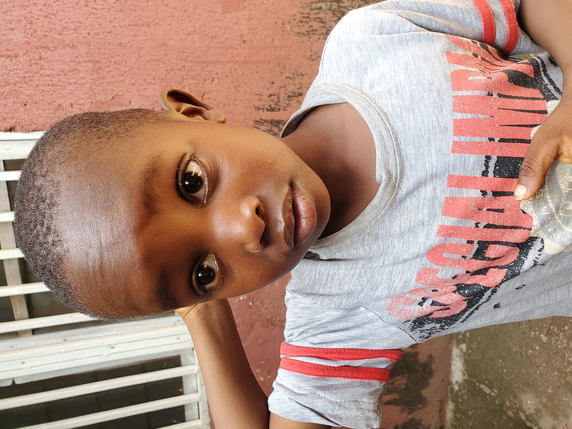 The image shows a close-up, rotated portrait of a young Black child resting their head on their arm. The child has dark skin, short curly hair, and is looking directly at the camera with a neutral expression. They are wearing a light grey t-shirt with red stripes on the sleeves and a distressed graphic print in red and black on the front. The background consists of a textured, reddish-brown wall and a white slatted structure, possibly a fence or window covering. The lighting suggests it is daytime, with a shadow cast across the child's forehead. The overall scene appears to be outdoors in a casual setting, likely in Jos, Nigeria, given the context. No discernible text is visible beyond the graphic on the shirt.