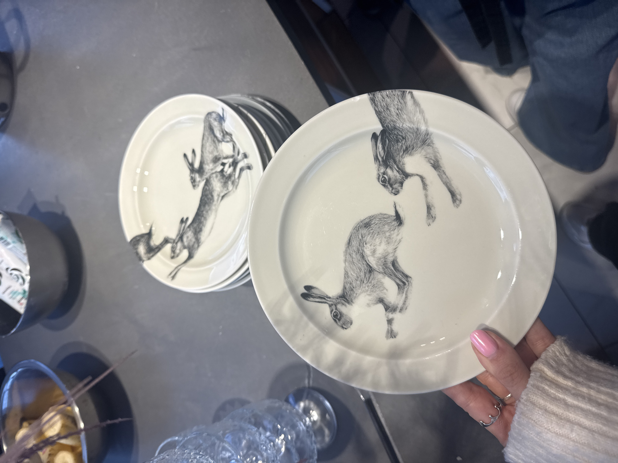 This indoor image, potentially taken in Izhevsk, Russia, focuses on a collection of white ceramic plates adorned with detailed black-and-white illustrations of hares.
In the foreground, a person's hand, featuring light pink nail polish and a silver ring, and clad in a light-colored ribbed sweater, holds one of these plates. On this particular plate, two hares are depicted: one appears to be leaping downwards from the top edge, while another is shown in an active, possibly rearing or jumping, posture. In the mid-ground, a stack of similar plates is visible, with the top plate clearly displaying a hare in a dynamic, upright stance.
The plates rest on a dark, smooth surface, likely a table or countertop. To the far left, a metallic bowl contains what looks like sliced bananas or similar light-colored fruit, accompanied by thin, dark decorative sticks. Above this, a dark cylindrical container holds white and green paper-like material, possibly packaging or napkins. A faceted glass object, perhaps a stemmed glass, is partially visible in the bottom center/right. In the upper right background, the lower half of a person's body, wearing dark blue jeans and light-colored footwear, is partially visible. The scene is illuminated by artificial lighting, creating some reflections on the glossy surfaces. No discernible text is visible anywhere in the image.