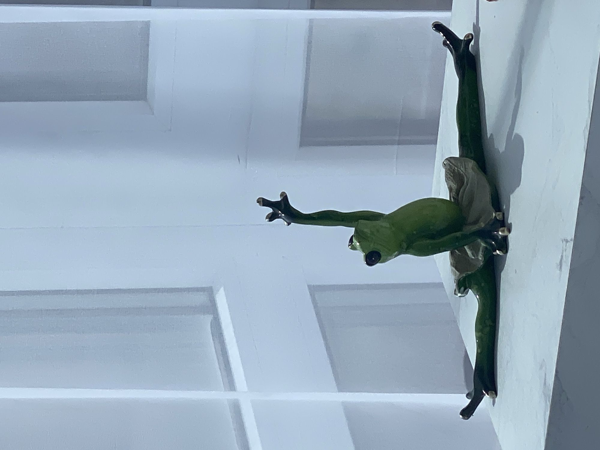 A small, green, ceramic frog figurine is positioned in the center of the frame, performing a ballet split. The frog is wearing a small, light-colored tutu. Its legs are extended horizontally to the sides, and its arms are raised, with one hand pointing upwards. The figurine is sitting on a flat, white surface that appears to be a table or pedestal. Behind the frog is a white, textured wall or backdrop with vertical lines, possibly representing a door or paneling. The lighting is soft and diffused, casting a slight shadow to the right of the frog. The overall scene is clean and minimalist, with the focus solely on the frog figurine.