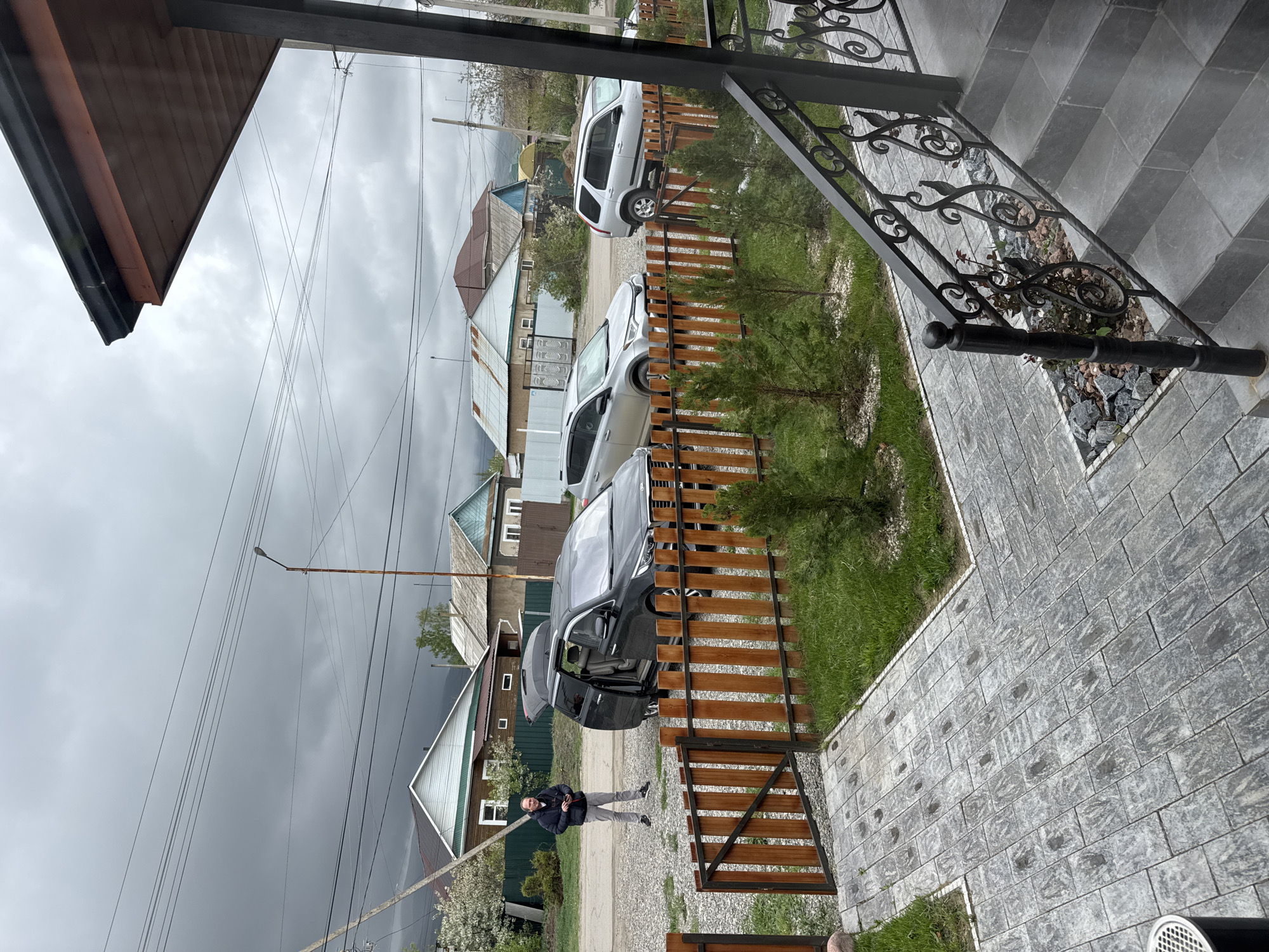 This image depicts a residential scene in Karakol City, Kyrgyzstan, captured on an overcast day. The perspective is slightly elevated, looking down onto a paved walkway and a street.
In the foreground, a dark gray paved walkway, composed of rectangular blocks, leads to a set of matching stairs on the right. A decorative black wrought iron railing with elaborate scrollwork borders the stairs and a small garden patch filled with rocks and sparse plants. To the left of the paved area, there's green grass and several small evergreen trees, separated by a wooden fence with dark metal frames.
In the midground, a man in a dark jacket and light pants stands on a gravel path, looking towards the camera with a neutral, slightly smiling expression. Behind him and to his right, a dark SUV or minivan is parked with its passenger-side doors and rear hatch open. Another silver SUV is parked further down the gravel path, behind the wooden fence.
The background reveals a residential neighborhood with several single-story and two-story houses featuring pitched roofs and various siding materials. Power lines crisscross the heavily overcast, dark gray sky, and a street lamp is visible. The heavy cloud cover suggests a cool, subdued atmosphere, possibly preceding rain. No legible text is visible in the image.