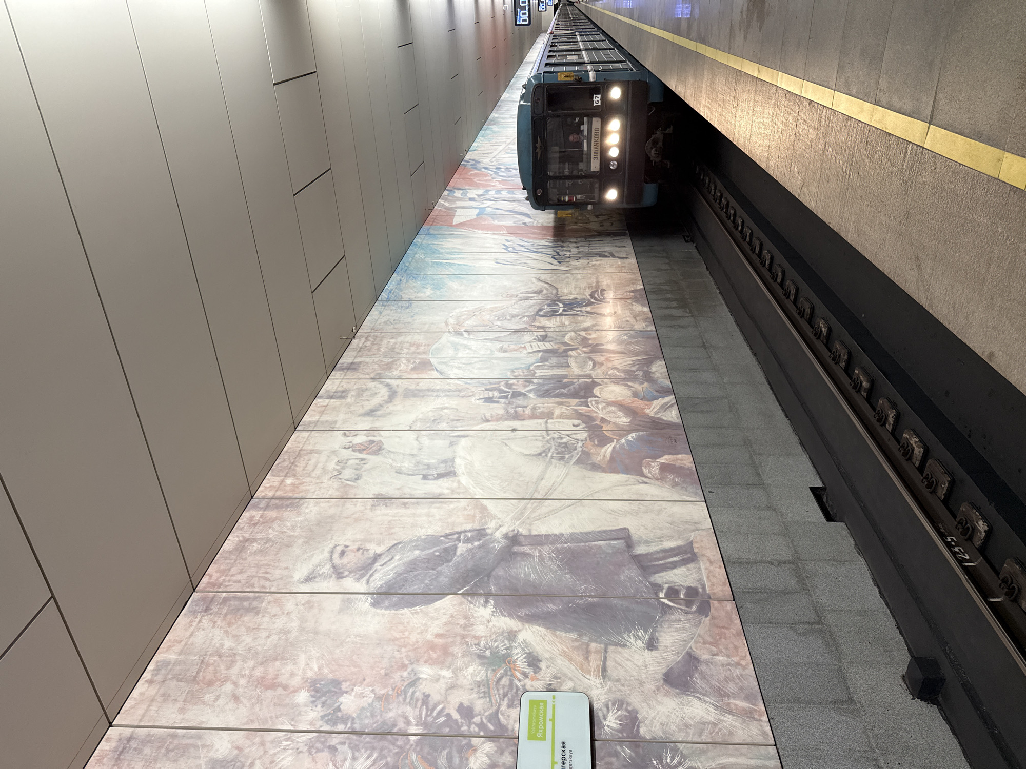 The image captures a subway station in Moscow, Russia, specifically the Yakhromskaya station, as indicated by the sign on the left. A teal-colored train is pulling into the station on the right side of the frame, with its headlights illuminating the platform. The station walls are adorned with a large, colorful mural that appears to depict historical scenes with figures in military attire, including a man on horseback in the foreground. The mural extends along the entire length of the platform visible in the image. The ceiling of the station is made of light-colored panels with metal seams. The platform itself is paved with grey tiles and marked with a bright yellow safety line along the edge. Train tracks run along the platform. The lighting in the station is functional, with overhead lights creating a well-lit environment. The overall atmosphere is that of a functional public transportation hub with artistic decoration.