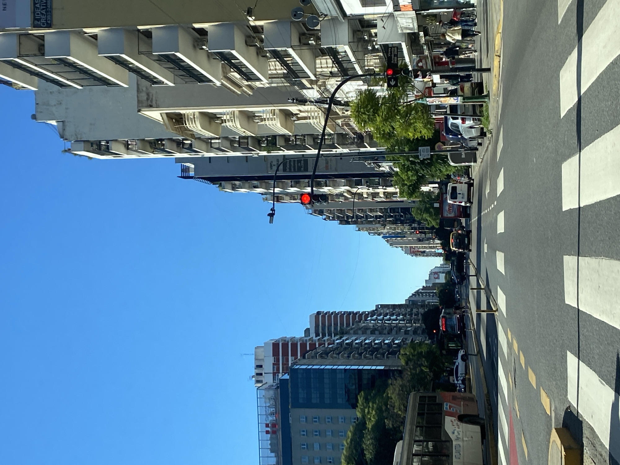 This image captures a vibrant street scene in Buenos Aires, Argentina, on a clear and sunny day. The perspective looks down a long, wide street flanked by numerous multi-story buildings on both sides, with a bright blue sky dominating the upper part of the frame.
The foreground and middle ground feature a paved road with distinct white pedestrian crosswalk stripes and yellow dashed lane markings. Several vehicles are visible, including cars, vans, and a white bus displaying "ATLANTIDA" on its side, indicating moderate traffic. Two red traffic lights are prominent, one closer to the viewer and another further down the street, suggesting traffic is currently stopped.
The buildings lining the street showcase varied urban architecture, ranging from structures with classic balconies and numerous windows to more modern facades, some with visible air conditioning units. Green trees are interspersed along the sidewalks, adding natural elements to the urban landscape. A few pedestrians can be seen walking on the sidewalks, engaging in typical daily city activities.
Visible text includes "IDEAS CREATIVAS" and "4381-1406" on a building facade, a partial "VISTA AL", and another sign advertising "Amoblamientos 24hs - 2506". The image depicts a bustling, active urban environment under bright daylight.
