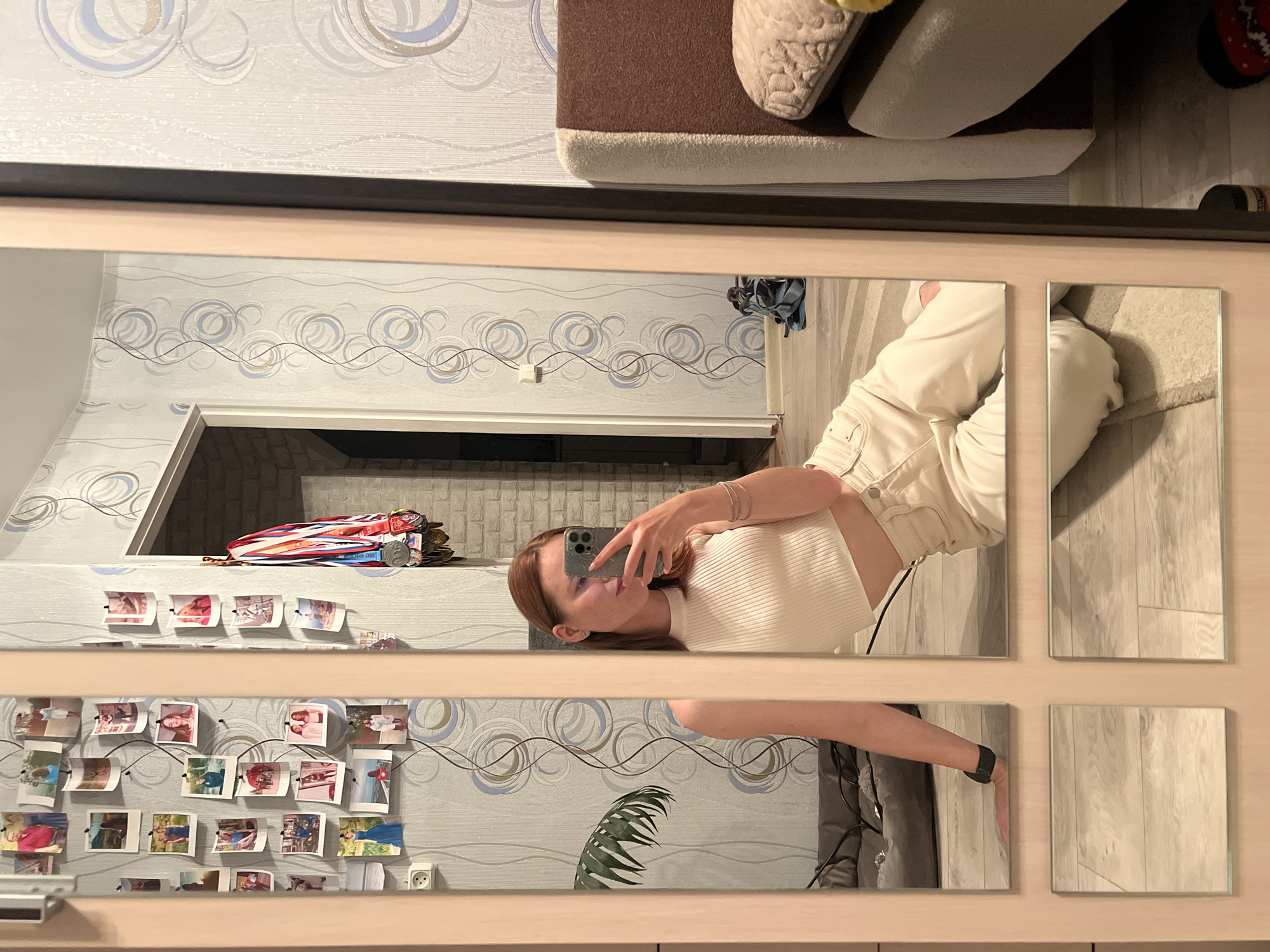 A young woman with reddish-brown hair is taking a mirror selfie in a room. She is wearing a white sleeveless crop top and cream-colored pants. She is sitting on the floor, leaning back on her left arm, with her right hand holding a smartphone to take the picture. The room has patterned wallpaper with circular designs and a light-colored wood-paneled doorframe. Several framed photographs are pinned to the wall on the left, and a string of medals hangs on the wall to the right. A brown and beige sofa is visible in the background on the right. The floor is made of light-colored wood laminate. The mirror itself is divided into multiple panels, creating reflections of the woman, the room, and a plant on the left. The image appears to be taken in a residential setting, possibly an apartment or house. The presence of medals suggests a potential athletic background. The overall atmosphere is casual and personal.