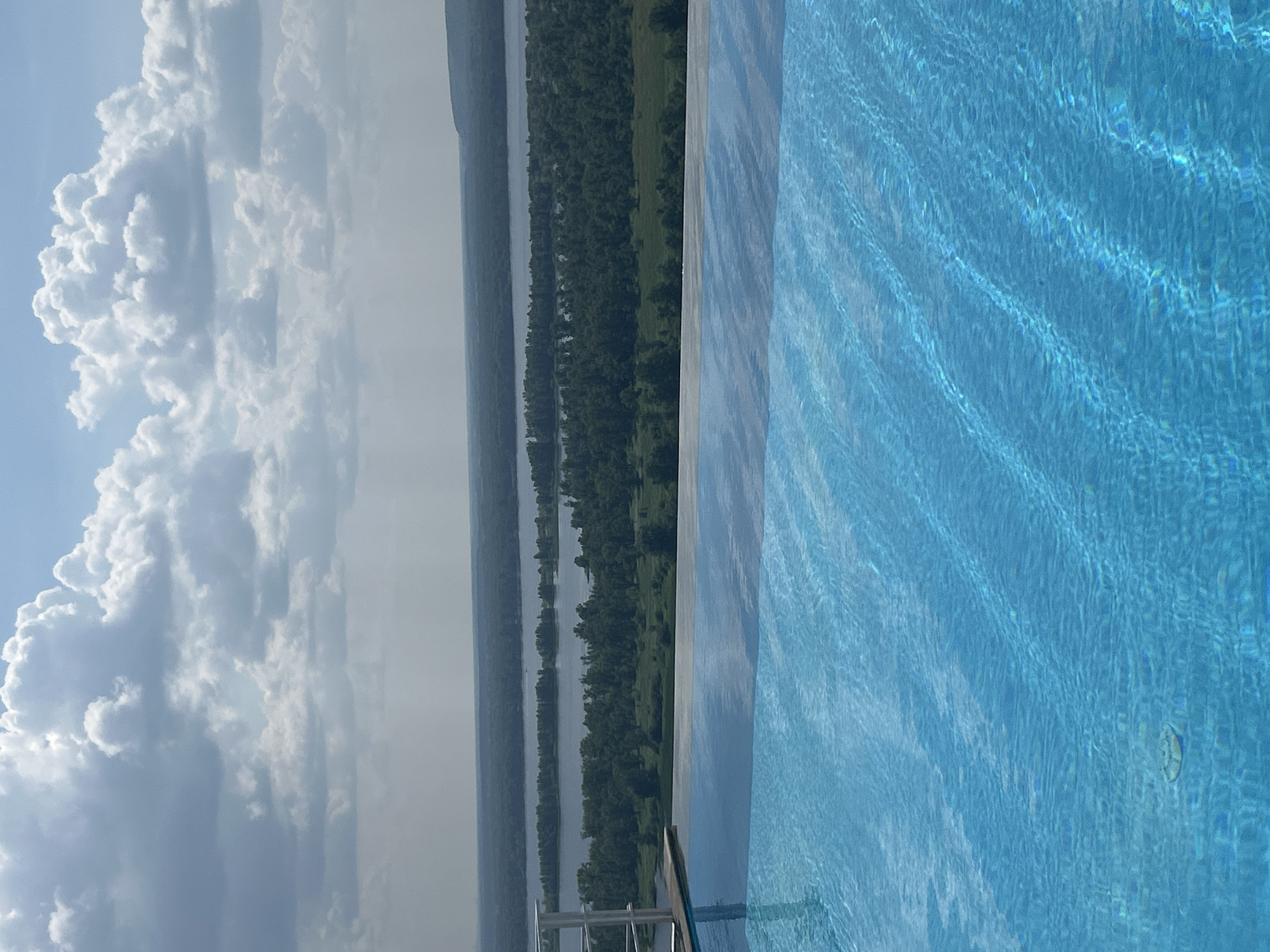 This image shows an infinity pool overlooking a wide river and a densely forested landscape under a cloudy sky. The pool's water is a bright blue with visible ripples and reflections. In the foreground, the edge of the pool and a glimpse of a wooden deck with a white railing are visible. The middle ground features the expansive river, bordered by lush green trees that stretch towards a hazy horizon. The sky is filled with dramatic, white and gray clouds. While the image depicts a serene natural setting, it is not possible to ascertain its specific location within Novokuybyshevsk, Russia, nor are there any people or specific actions depicted.