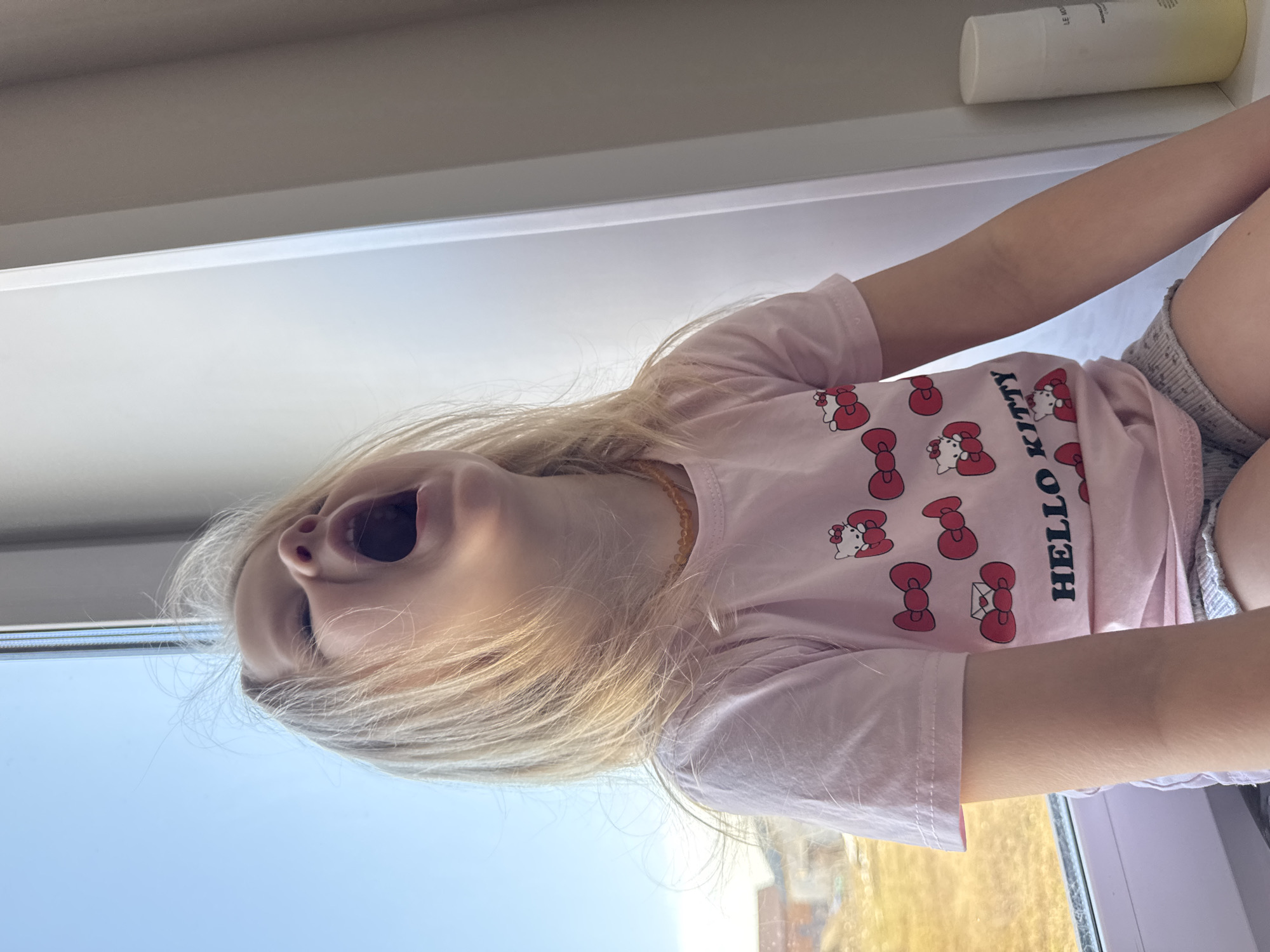 A young child with blonde hair, wearing a pink Hello Kitty t-shirt and grey shorts, is positioned in front of a window. The child's mouth is open. The view outside the window is blurred, suggesting a landscape. A white bottle sits on the window sill. The image may be taken in the city of Gornyi (Dvurechenskoe S/P), Russia.