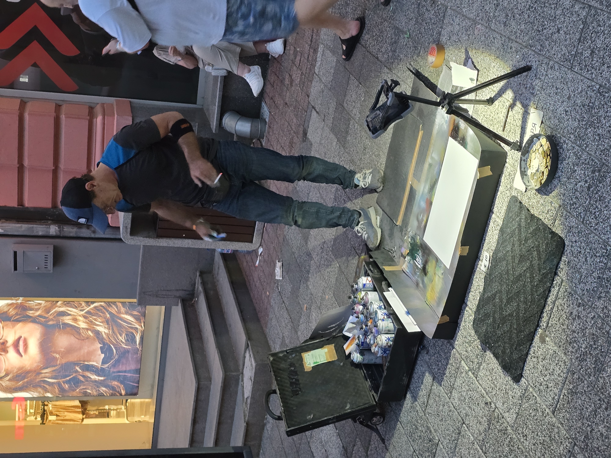 The image depicts an outdoor street scene in a pedestrian area, identified as Plovdiv, Bulgaria. In the foreground and center, a male street artist is standing, dressed in a blue and black baseball cap, a dark grey t-shirt with blue shoulder panels, blue jeans, and light-colored sneakers. He wears a black fanny pack and a black armband on his left arm. His gaze is directed downwards towards his art setup.
To the artist's left, a large, open black hard-shell case on wheels rests on the grey cobblestone pavement. The case is filled with numerous spray paint cans of various colors, including white, silver, blue, purple, pink, and yellow. Adjacent to the case, a flat, dark rectangular panel serves as a workspace, holding a blank white sheet of paper and showing traces of multi-colored paint. A small, black rectangular mat is placed on the ground in front of this workspace, with a small white sign displaying "€ 10" next to it.
To the artist's right, a black tripod stand supports a light source, which illuminates the workspace and a black bowl filled with gold-colored coins, placed directly on the pavement. A piece of paper with partial Cyrillic text, reading "ря", is visible next to the money bowl. A roll of brown packing tape and other small paper scraps are scattered on the ground nearby.
In the background, behind the artist, a building facade features a light pink/red brick section with a dark shop window displaying red, arrow-like graphics. To the left, another shopfront has a large poster of a woman with long, wavy, light brown hair and glasses. The shop interior, displaying clothing, is visible through the window. A set of grey stone steps leads up to this shop entrance. A grey metal mailbox is affixed to the wall between the two shopfronts. Further to the right, a metal pipe protrudes from the building wall.
Several other individuals are partially visible in the scene. On the far right, a person in a white t-shirt and patterned shorts, wearing dark flip-flops, is walking past. On a raised ledge behind the artist, two people are seated; one is wearing light-colored trousers and white sneakers. The overall lighting suggests late afternoon or early evening, with a mix of natural light and artificial illumination from shop windows and the artist's dedicated light.